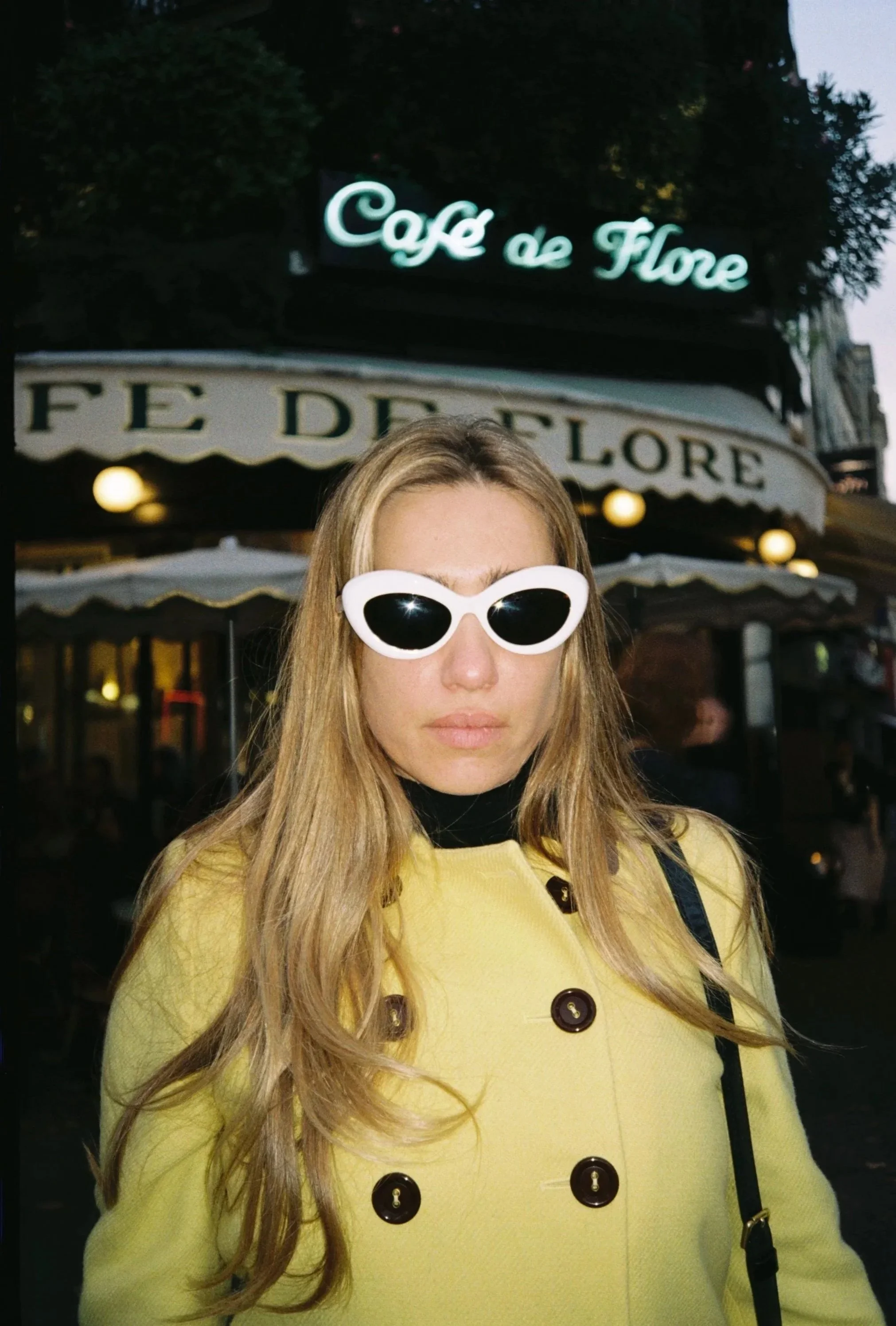 A woman with long blonde hair wearing white sunglasses and a yellow coat standing in front of the Cafe de Flore sign.