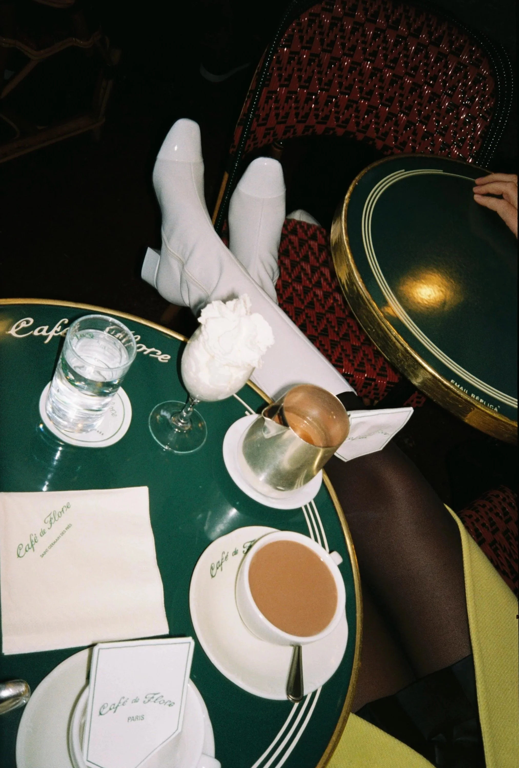 A table at Café de Flore with coffee, water, and dessert, and a person sitting with crossed legs in white boots and patterned clothing.