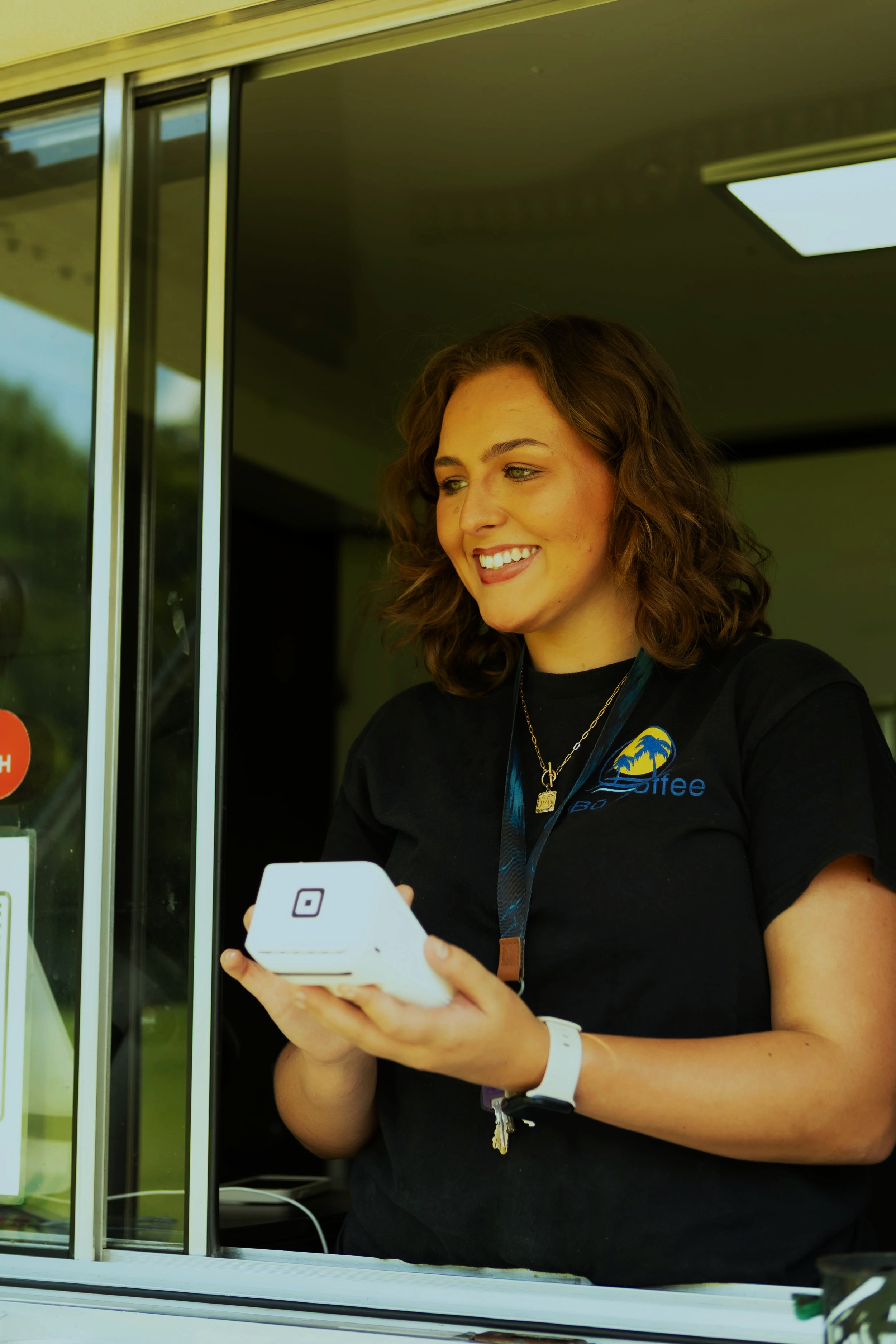 A young woman with curly brown hair and a smile is looking at her phone while working at a drive-thru window. She is wearing a black uniform with a logo on her shirt and a white wristwatch.