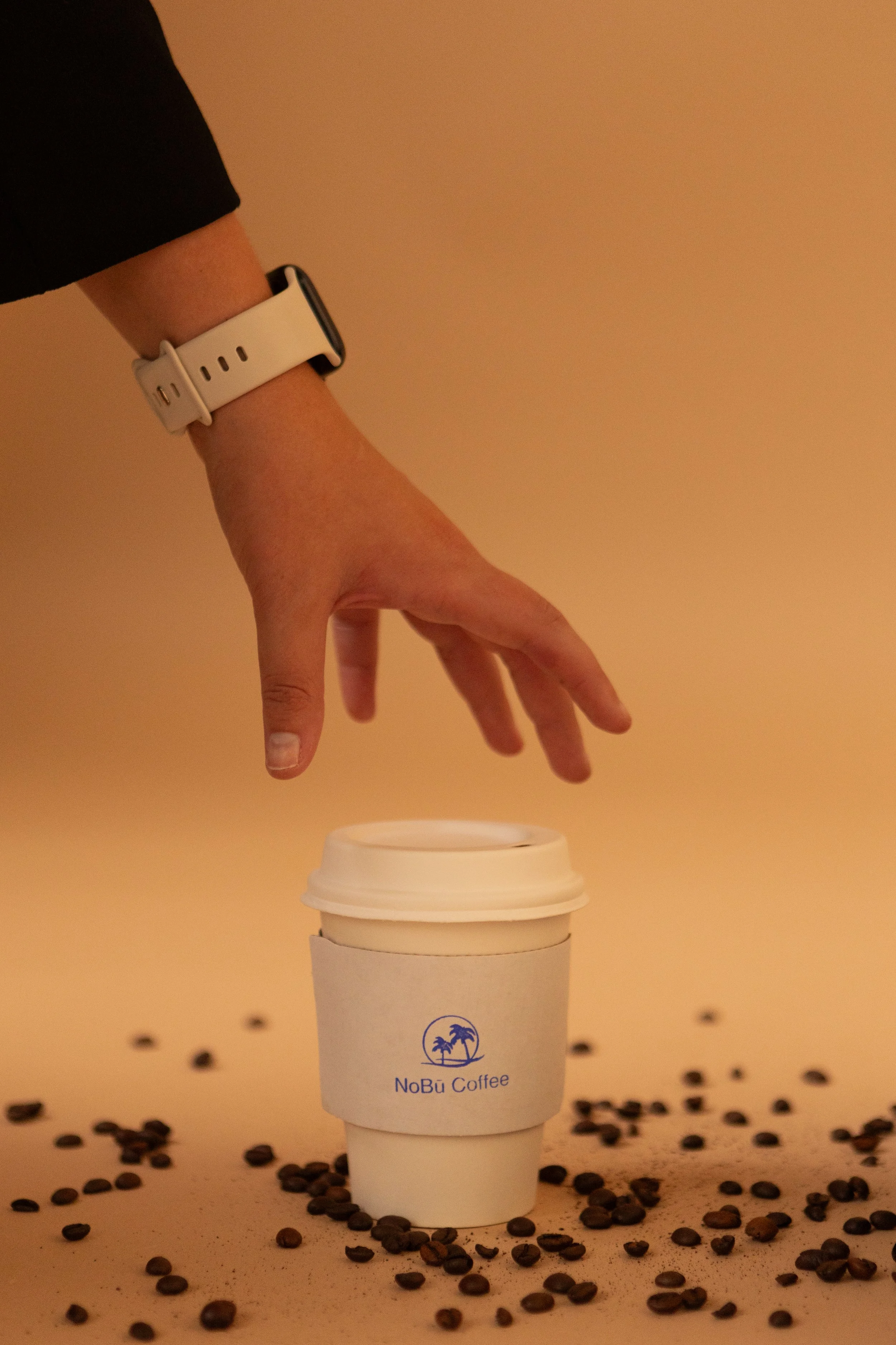 A hand with a white smartwatch reaching for a to-go coffee cup with a NoBu Coffee logo, surrounded by scattered coffee beans on a beige surface.