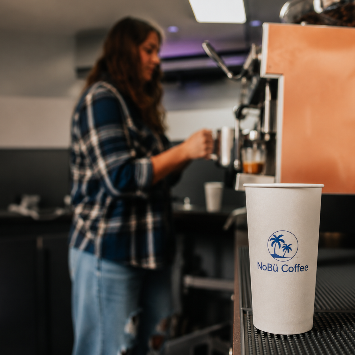 A paper coffee cup with NoBu Coffee logo on a counter, with a woman in a blue plaid shirt and jeans making coffee in the background.