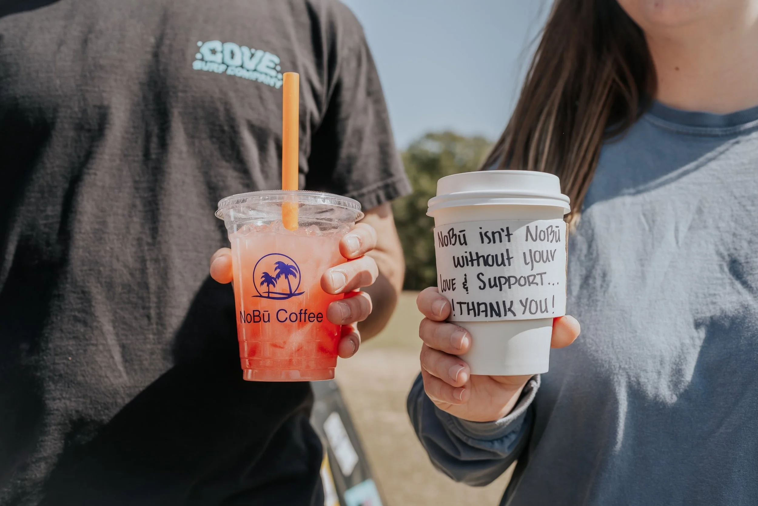 Two people holding beverages, one with a pink frozen drink in a clear cup with a straw, and the other with a white disposable cup with a handwritten thank you message. The person with the frozen drink wears a dark gray shirt with light blue lettering, and the other person wears a light blue shirt with long brown hair.