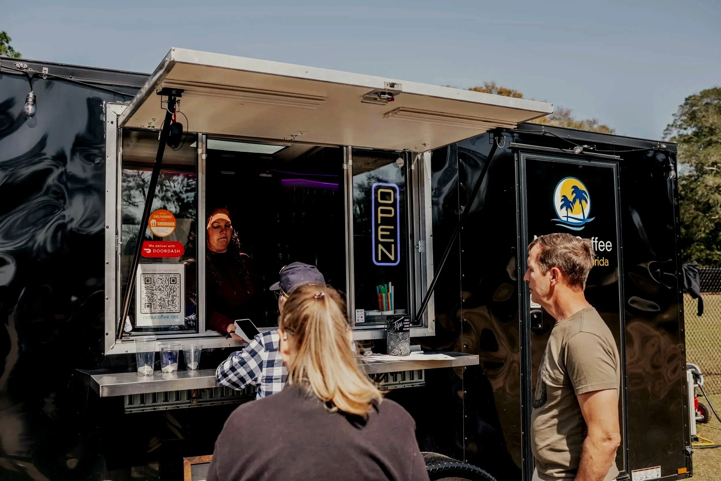 A black coffee truck with a window open, serving customers outdoors. Several people are gathered in front, including a woman with long curly hair inside the truck, a man in a gray shirt, and others with their backs turned. The truck has signage with a palm tree logo and a QR code for a website.