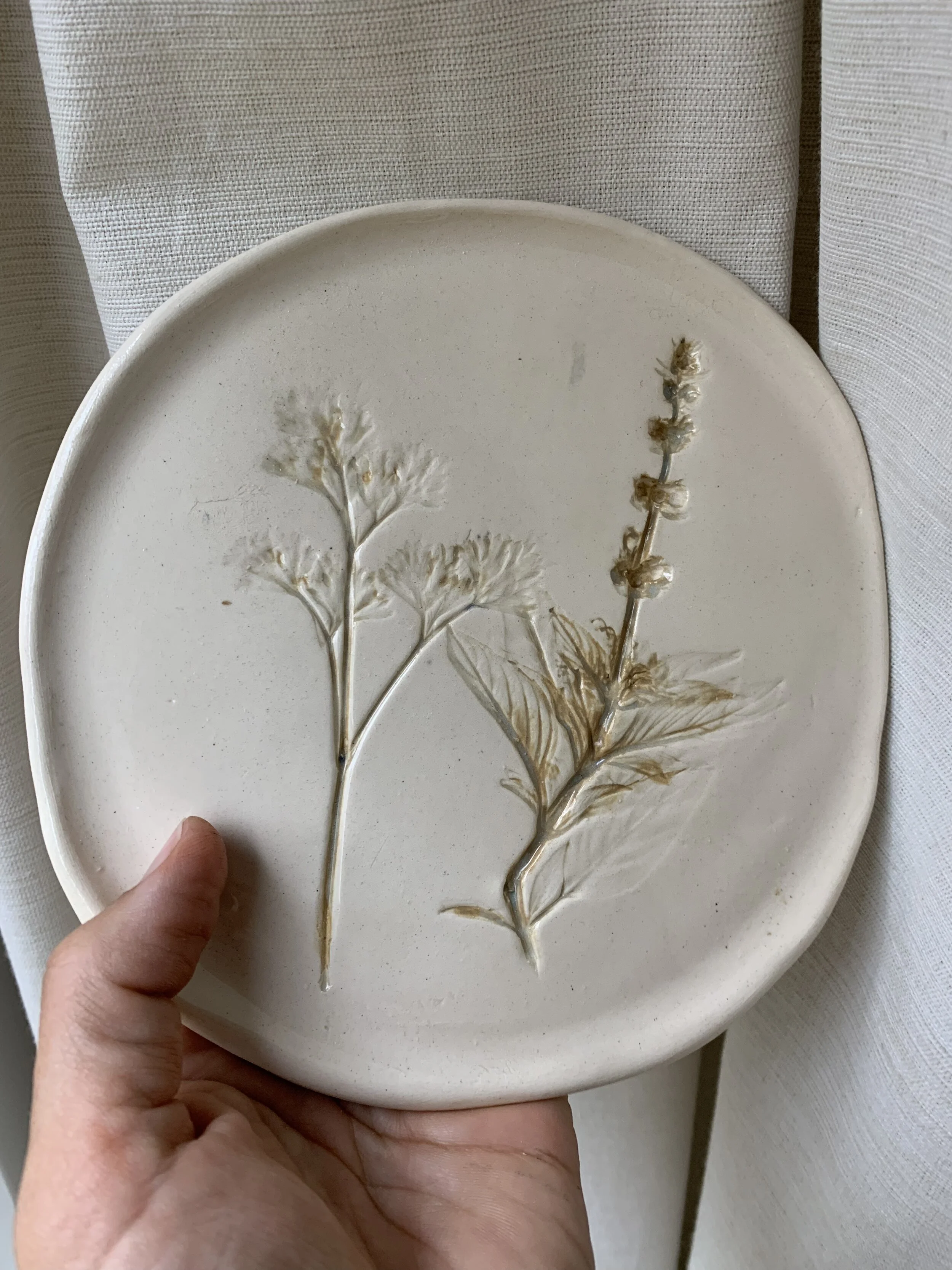 Ceramic plate with embossed plant designs held in a person's hand, background of white textured fabric.