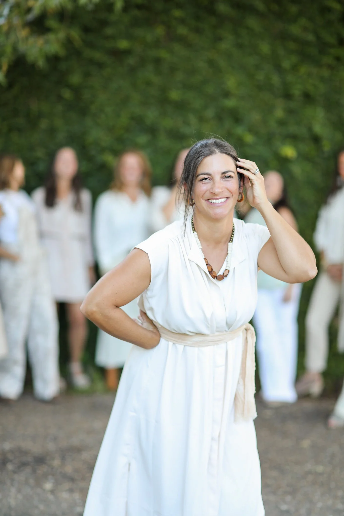 A woman in a white dress smiling and touching her hair, standing outdoors with a green leafy background and several other women in white outfits blurred in the background.