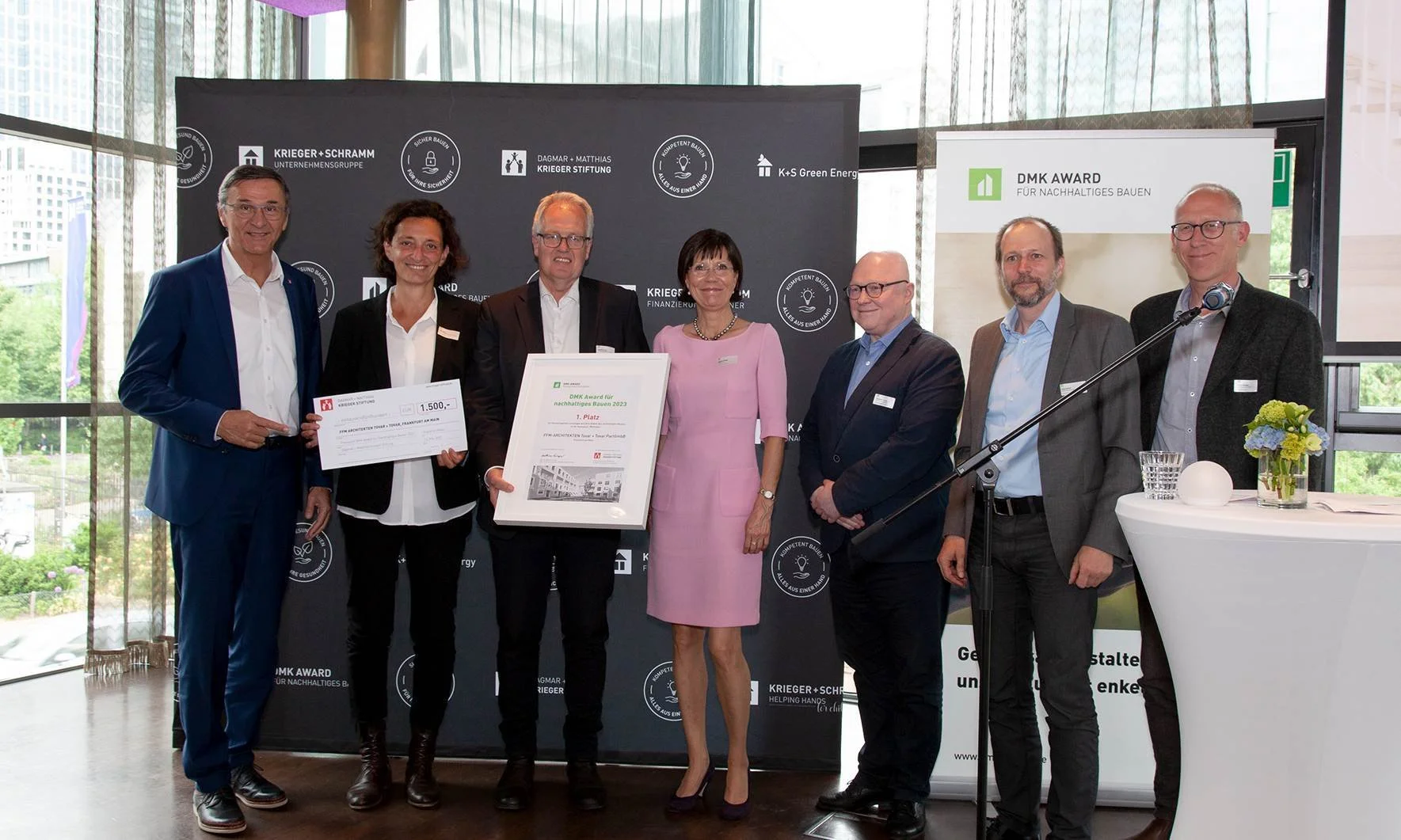 Eine Gruppe von sieben Personen posiert bei einer Preisverleihung. Zwei Personen halten ein Zertifikat und einen Scheck über 1.500 Euro. Im Hintergrund sind Sponsorenlogos und ein Banner mit der Aufschrift 'DMK Award für nachhaltiges Bauen'. Die Personen sind formell gekleidet.