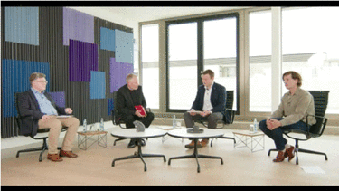 Vier Männer sitzen in einem modernen Konferenzraum auf Stühlen um einen Tisch herum. Der Raum hat große Fenster und schallisolierende Paneele an der Wand.