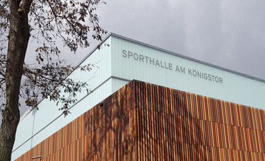 Gebäude mit der Aufschrift 'Sporthalle am Königstor' und einem Baum im Vordergrund.
