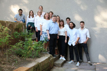 Gruppe von Personen steht auf einer Treppe im Freien, lächelnd in die Kamera schauend, vor einer grauen Wand und grünem Hintergrund.