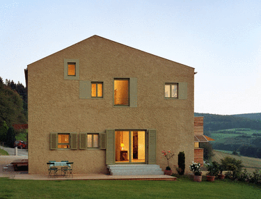 Modernes Haus mit Garten in ländlicher Umgebung, große Fenster, beleuchtete Innenräume, Wiese im Vordergrund und Hügel im Hintergrund.