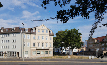 Straßenszene mit historischen Gebäuden, Bäumen und Laternen in einer städtischen Umgebung.