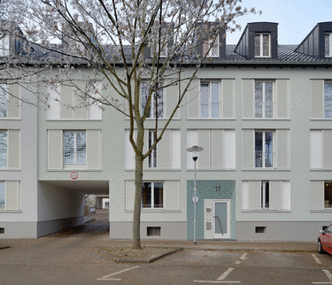 Wohngebäude mit moderner Architektur, blauer Eingangstür und großen Fenstern; Baum und beleuchtete Straßenlaterne im Vordergrund.