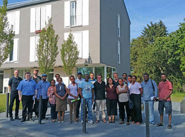 Eine Gruppe von Menschen steht vor einem modernen, mehrstöckigen Gebäude bei sonnigem Wetter. Es gibt Bäume im Hintergrund und Pflastersteine auf dem Boden. Die Personen sind in lässiger Kleidung und lächeln in die Kamera.