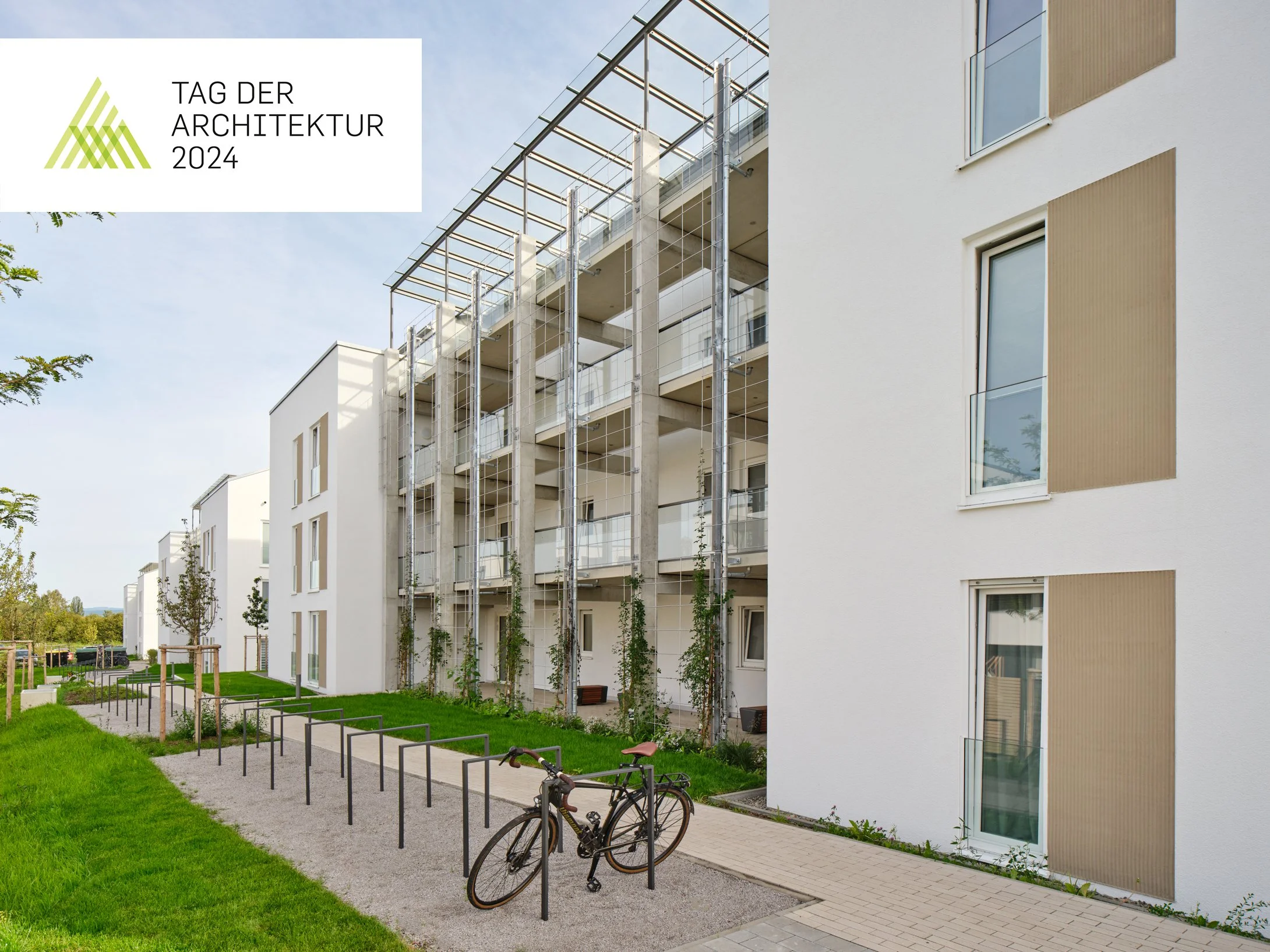 Moderne Wohngebäude mit Balkon, Fahrradständer im Vordergrund, grüner Rasen und Gehweg, Tag der Architektur 2024 Logo in der Ecke.