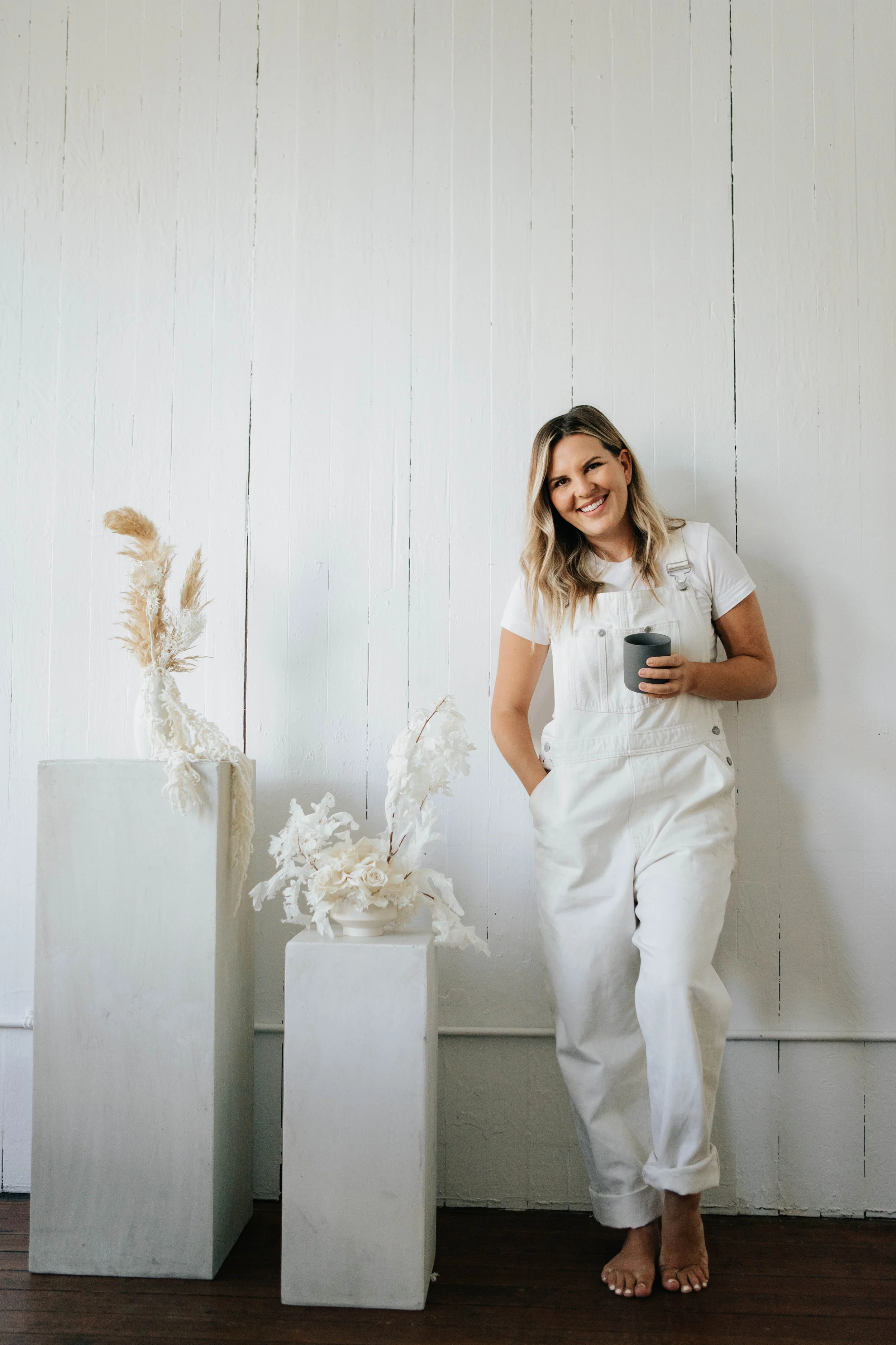 A woman in white overalls smiling and holding a black mug in a minimalist room with white wooden wall and white vases with dried flowers.