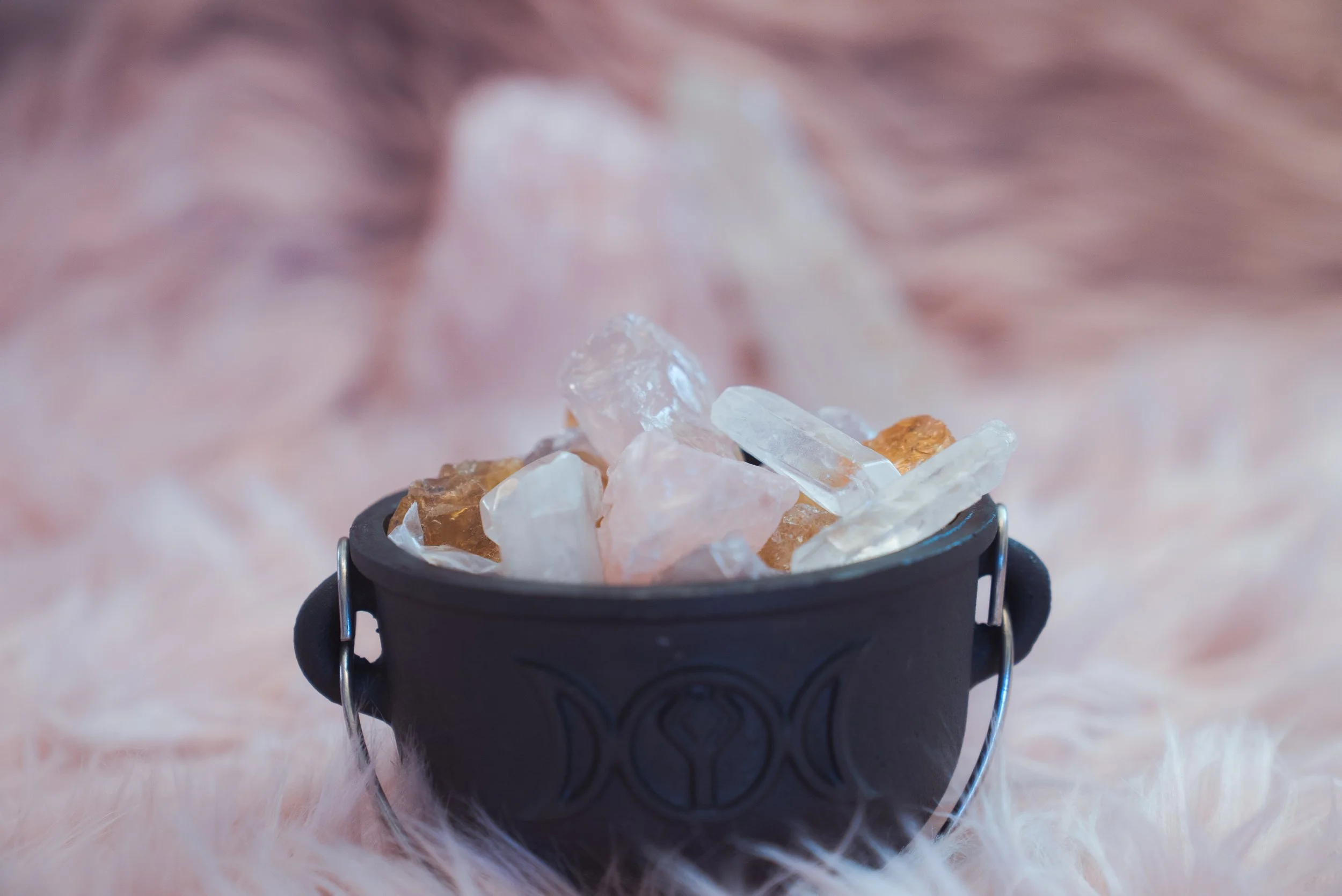 A small black cauldron filled with clear and amber-colored crystals, placed on soft pink fur.