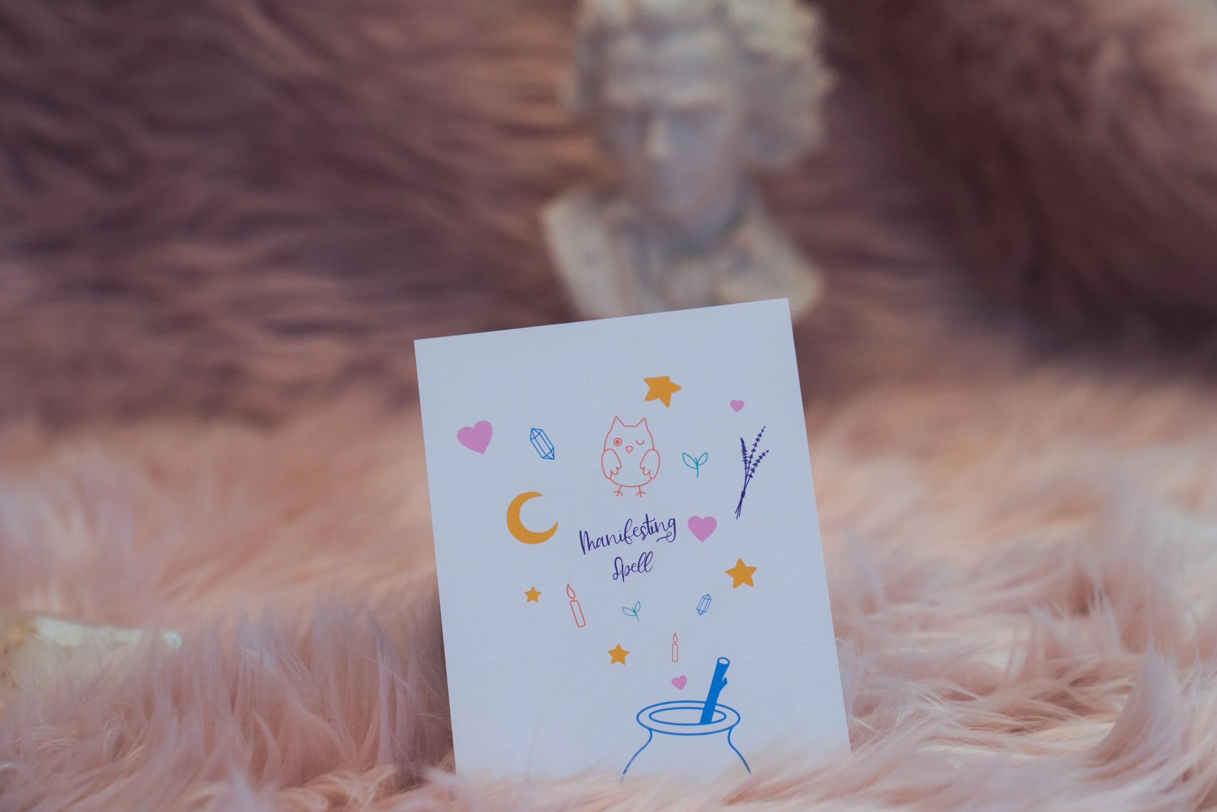 A card with colorful illustrations of hearts, stars, candles, and an owl, with the words "Manifesting Spell" written in purple, placed on a soft pink fluffy surface, with a blurred background.
