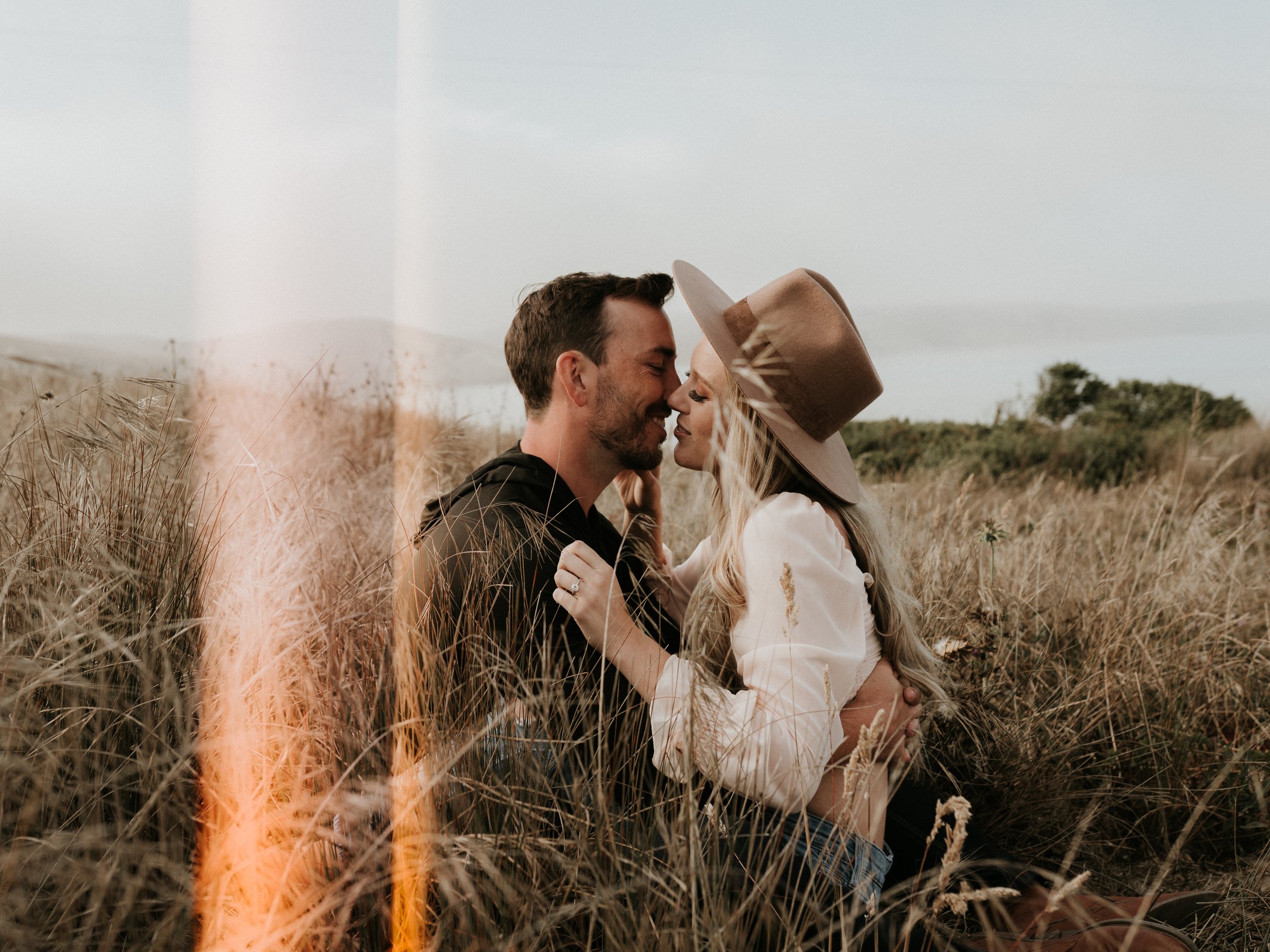 Couple embracing and smiling in a grassy field, woman wearing a hat.