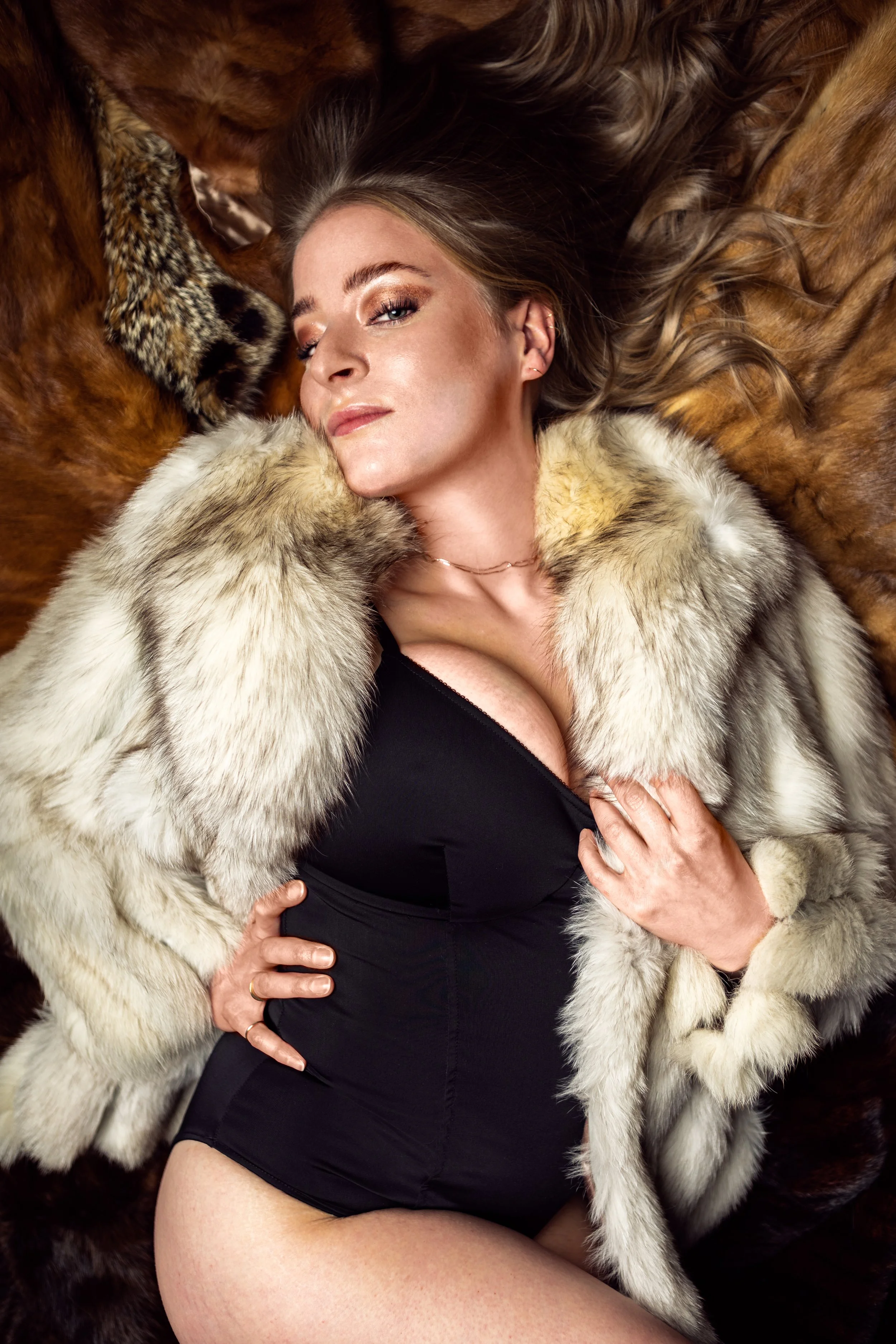 A woman with makeup and long hair lying on a surface covered with various animal furs, wearing a black outfit and a fur stole.