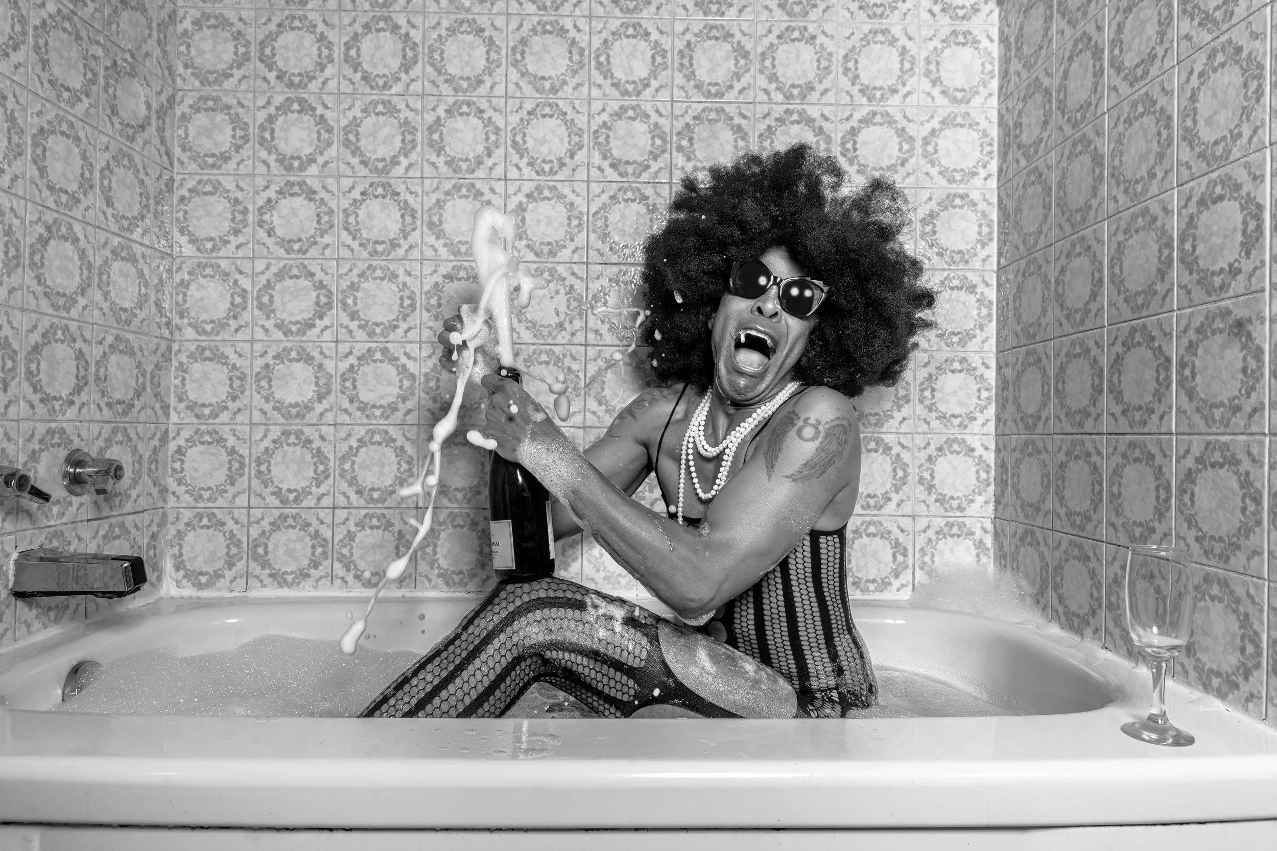 A woman with curly hair, wearing sunglasses, pearl necklaces, and striped pants, sitting in a bathtub with soap bubbles, holding a champagne bottle, and appearing surprised or excited. There is a glass of wine on the edge of the tub.