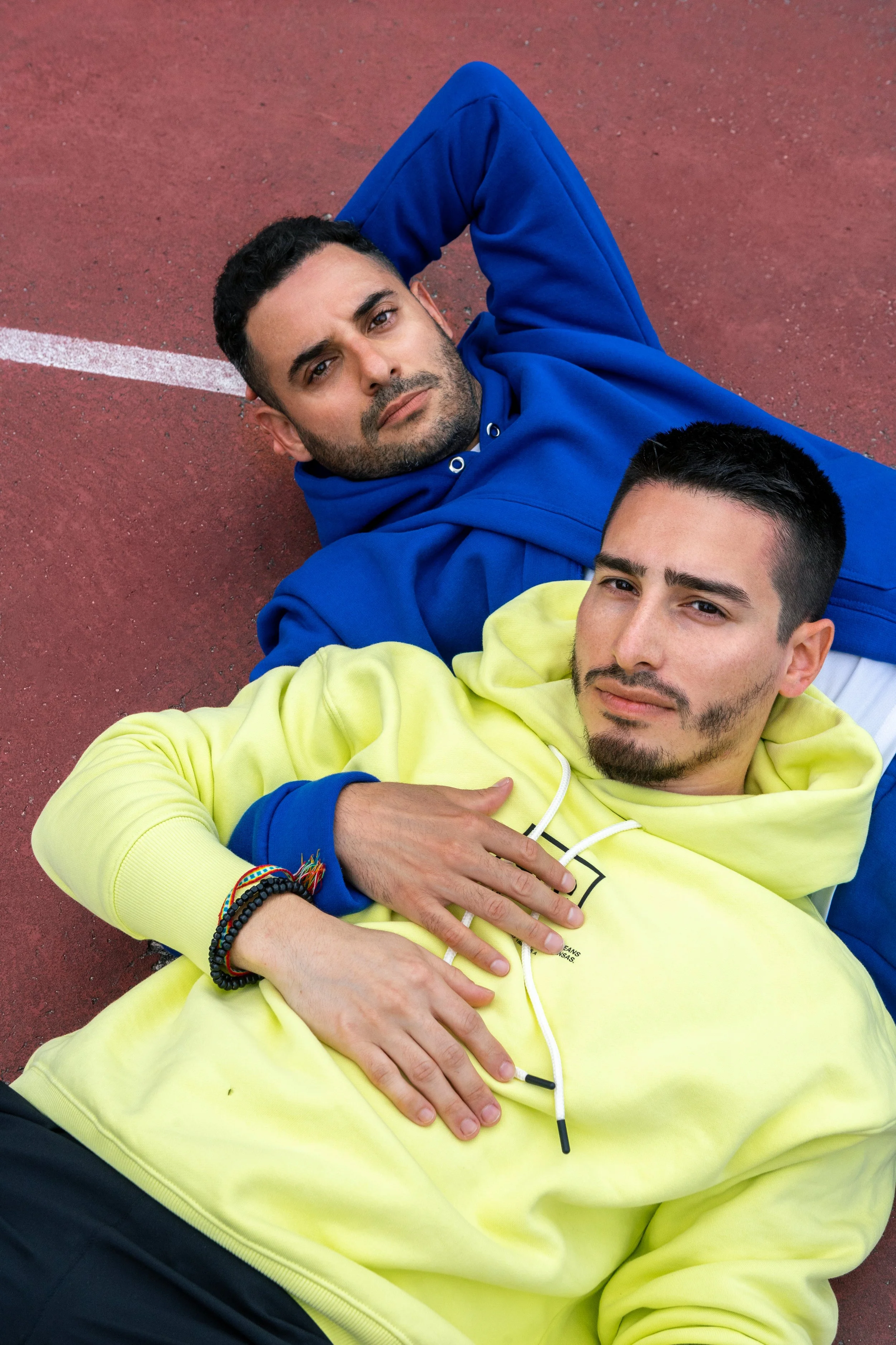 Two young men lying on a red outdoor sports surface, one wearing a blue hoodie and the other in a yellow hoodie, looking into the camera.