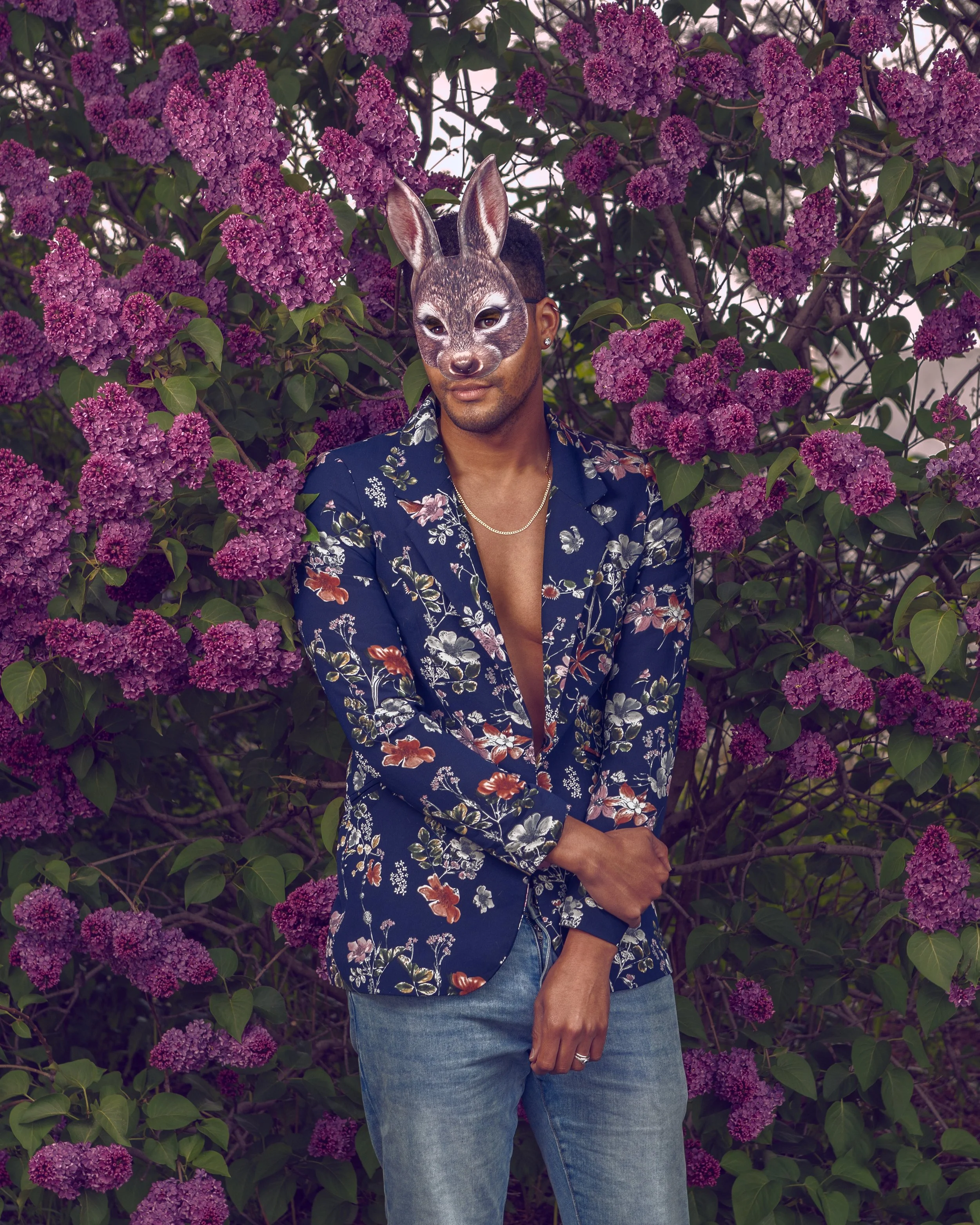 Person standing in front of a bush with purple flowers, wearing a floral jacket, jeans, and a rabbit mask over their face.