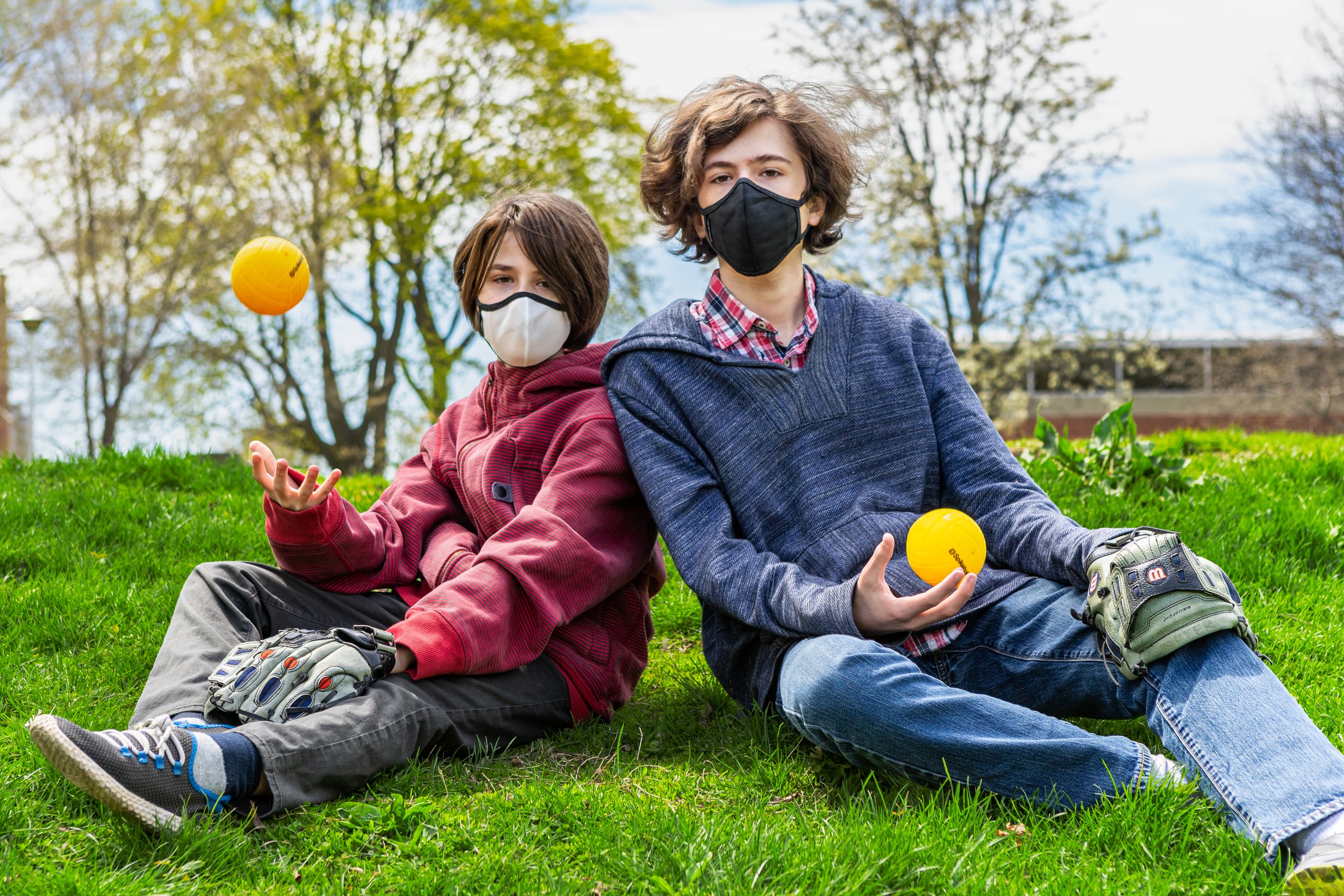 Two boys sitting on grass playing with yellow balls, wearing face masks, with trees in the background.
