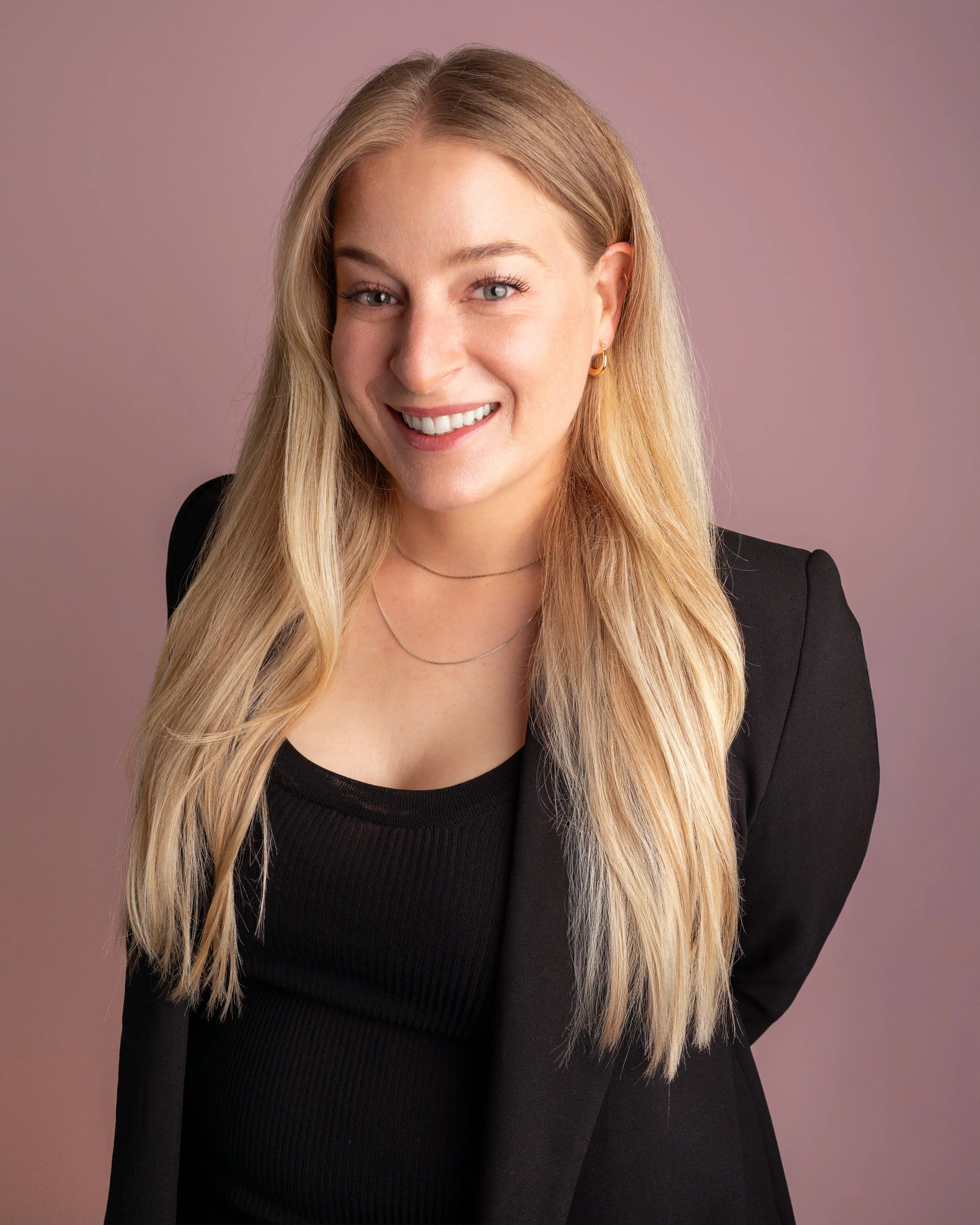 Portrait of a smiling woman with long blonde hair, wearing a black blazer and black top, against a soft pink background.