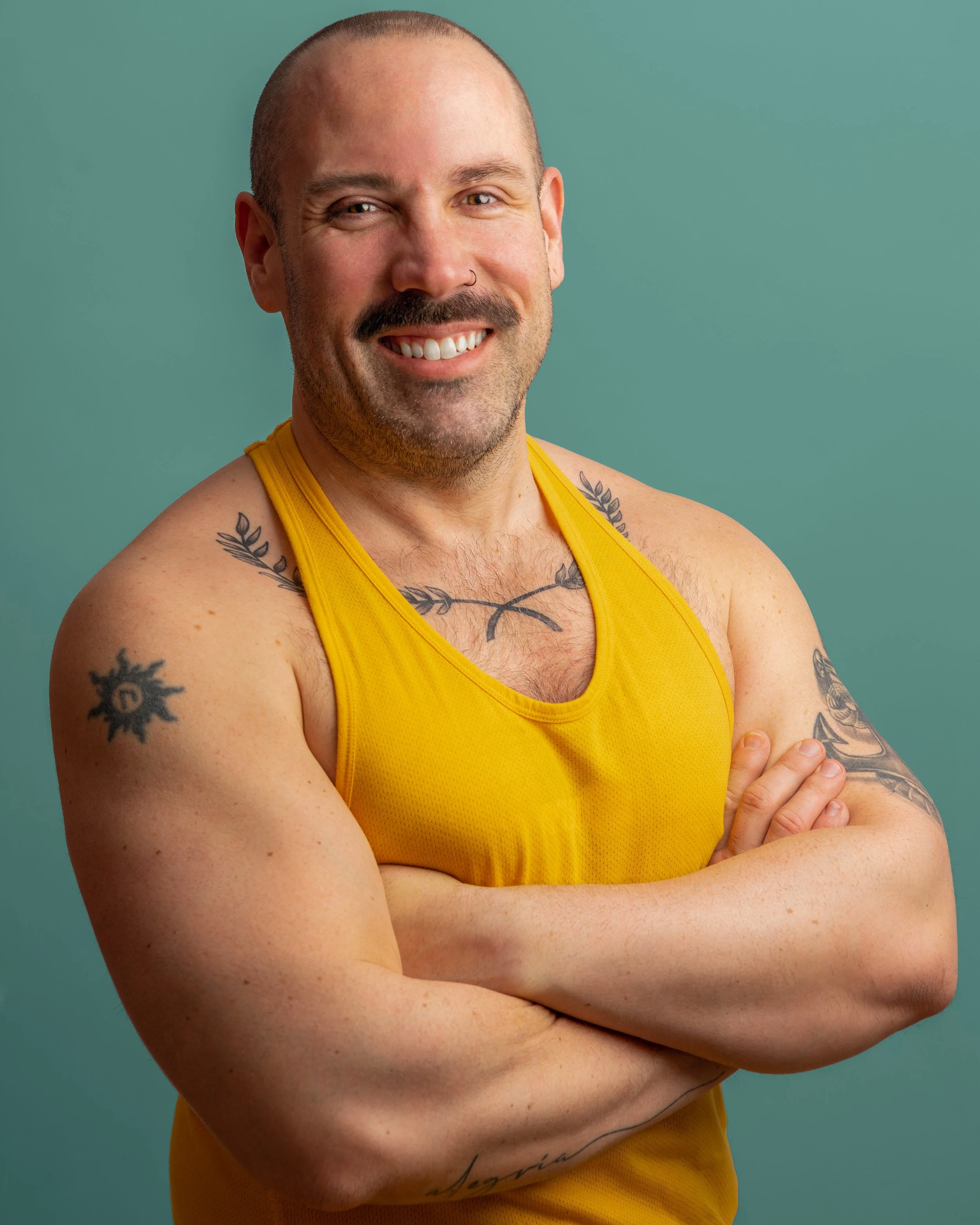 A smiling man with tattoos on his arms and chest, wearing a yellow sleeveless shirt, standing with arms crossed against a teal background.