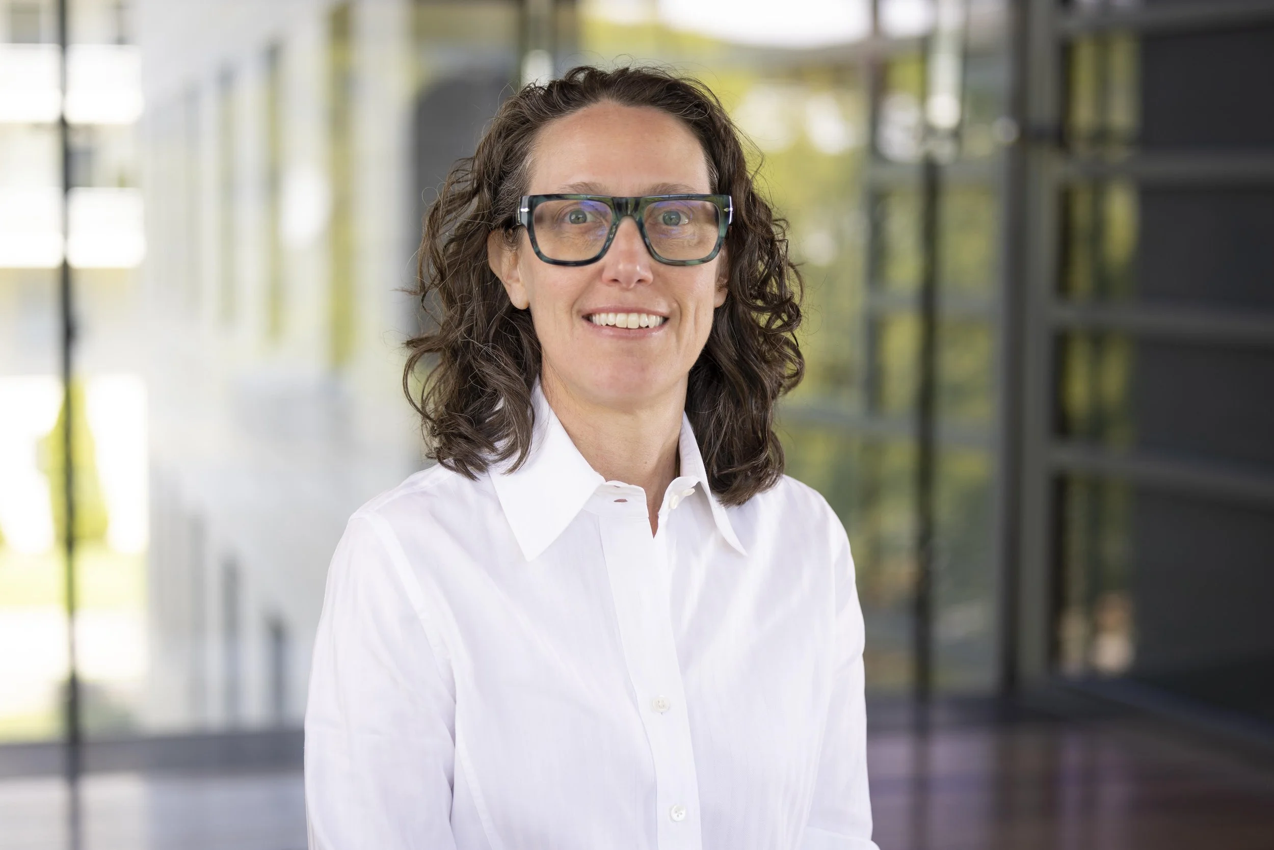 A woman with curly brown hair, wearing black glasses and a white button-up shirt, smiling with her arms crossed.