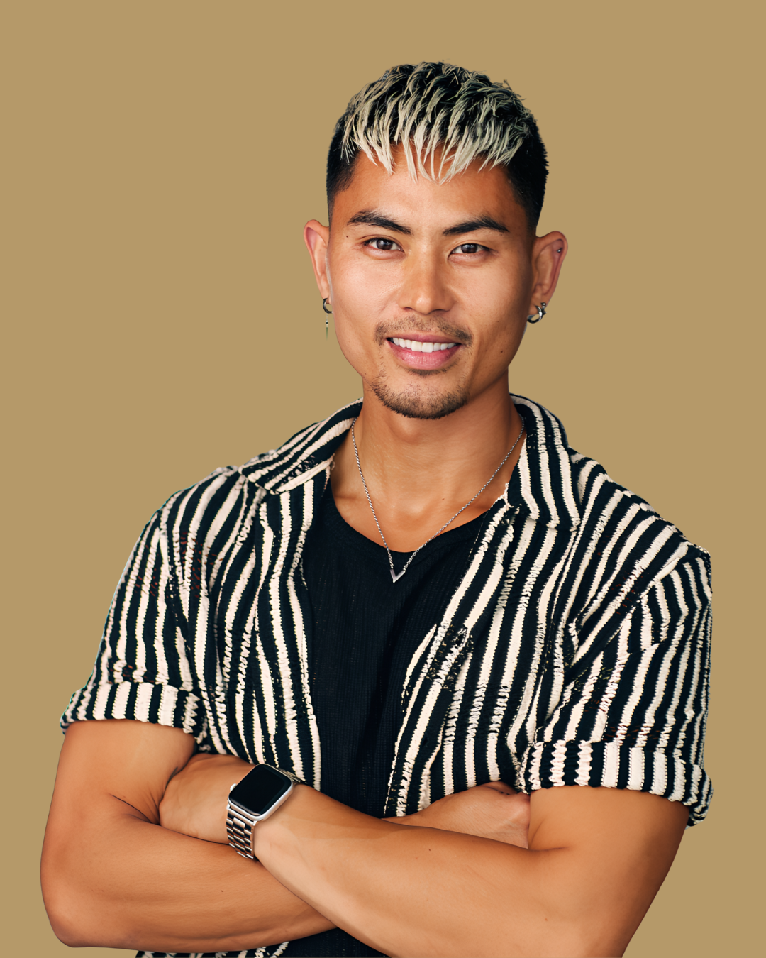 A young Asian man with short, dyed blonde hair, wearing a black shirt and a striped short-sleeve shirt layered over it, with earrings, a necklace, and a smartwatch, smiling with arms crossed against a beige background.