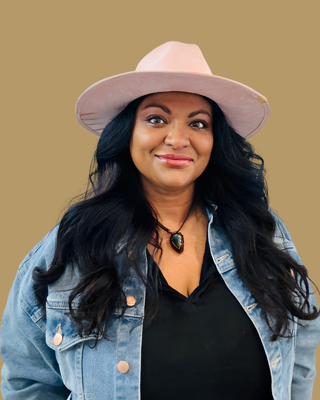 A woman with long dark hair wearing a pink wide-brimmed hat, a black top, a denim jacket, and a necklace with a green pendant, smiling against a tan background.