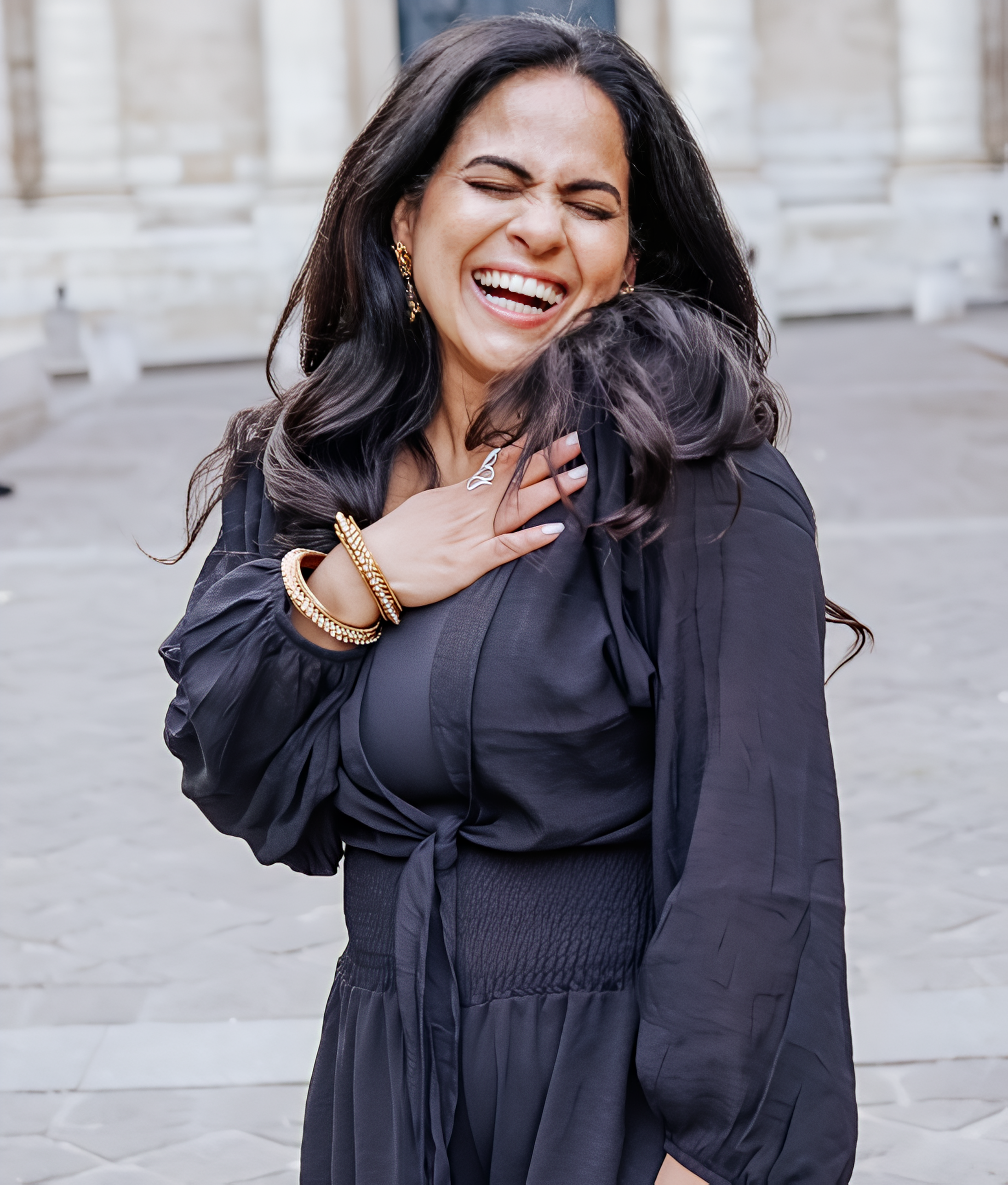 Woman in black dress laughing outdoors with hand on chest