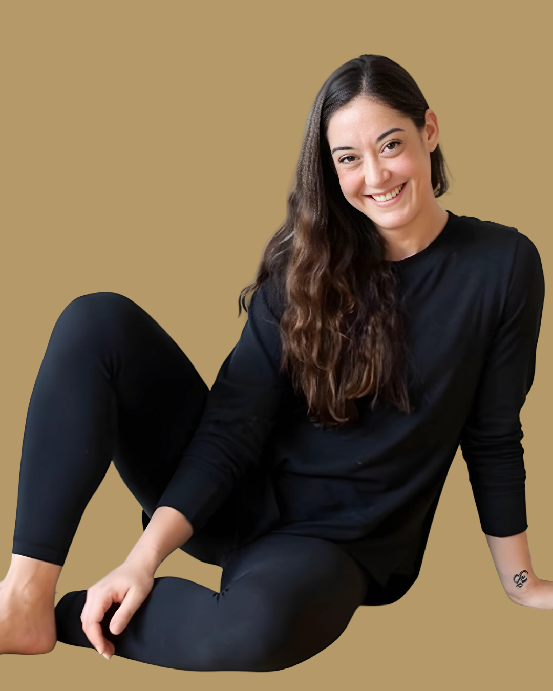A woman with long brown wavy hair smiling, wearing a black long-sleeve top and black pants, sitting against a plain beige background.