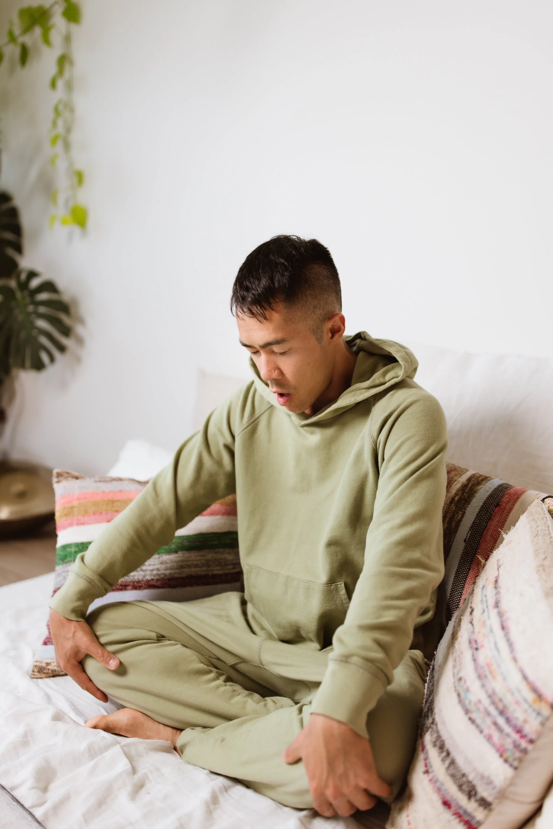 Person in green hoodie meditating on a bed with colorful cushions.