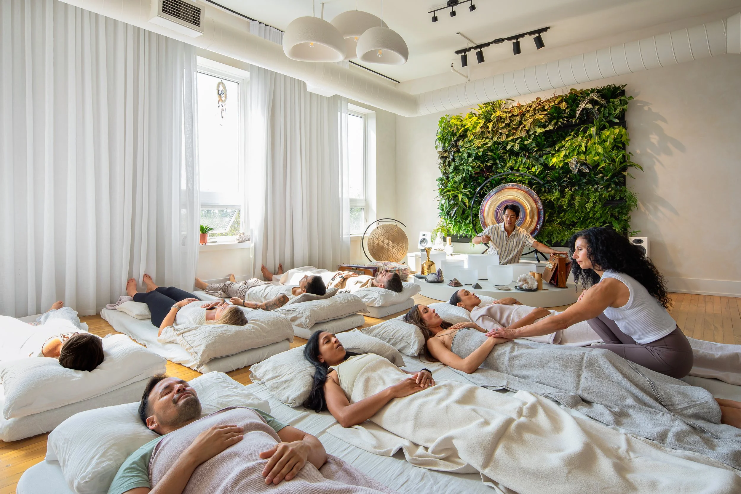 Group of people lying on mats in a peaceful room during a sound bath meditation, with a practitioner playing instruments near a green wall.