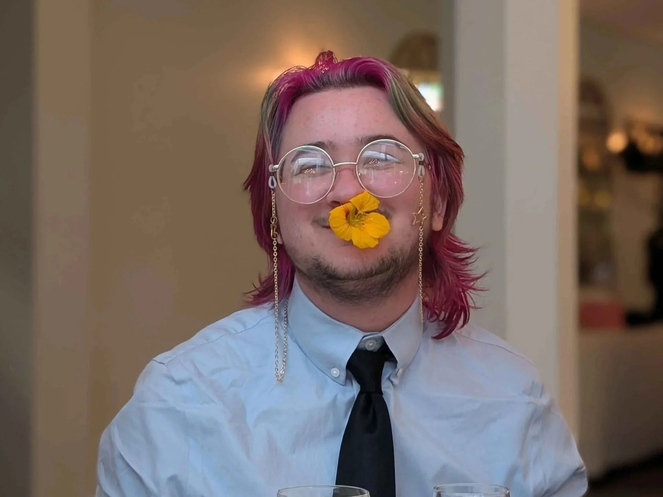 A person with pink and green hair wearing glasses, a light blue shirt, and a black tie, holding a yellow flower in their mouth, indoors with blurred background.
