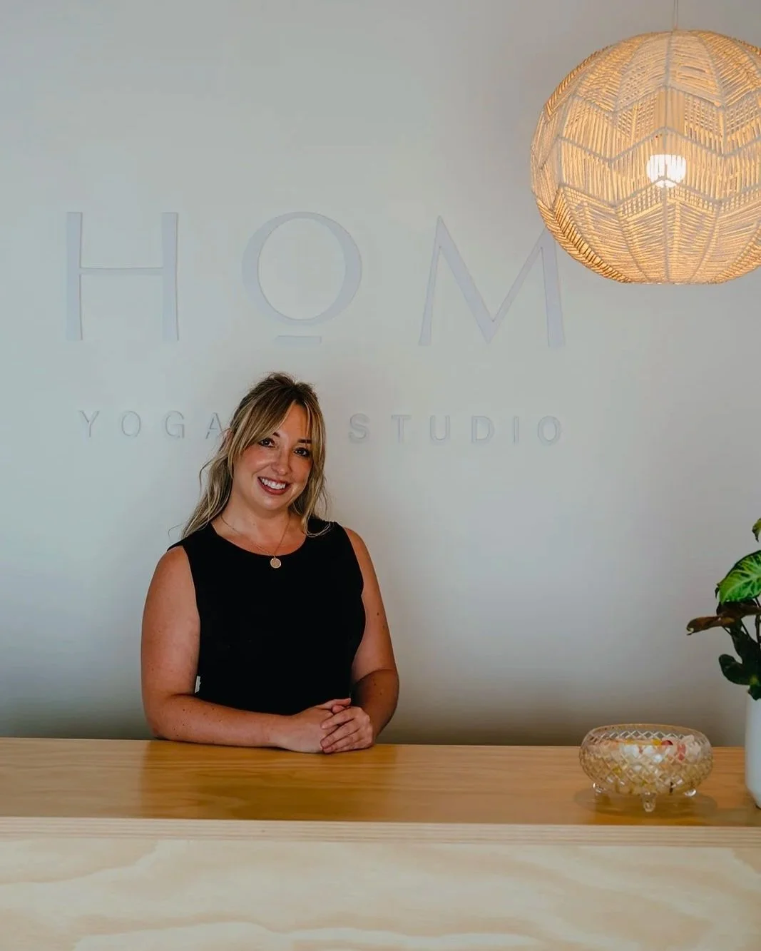 Woman smiling at a yoga studio reception desk with a sign reading 'HOME YOGA STUDIO' in the background, a woven pendant light overhead, and a plant on the right side of the desk.