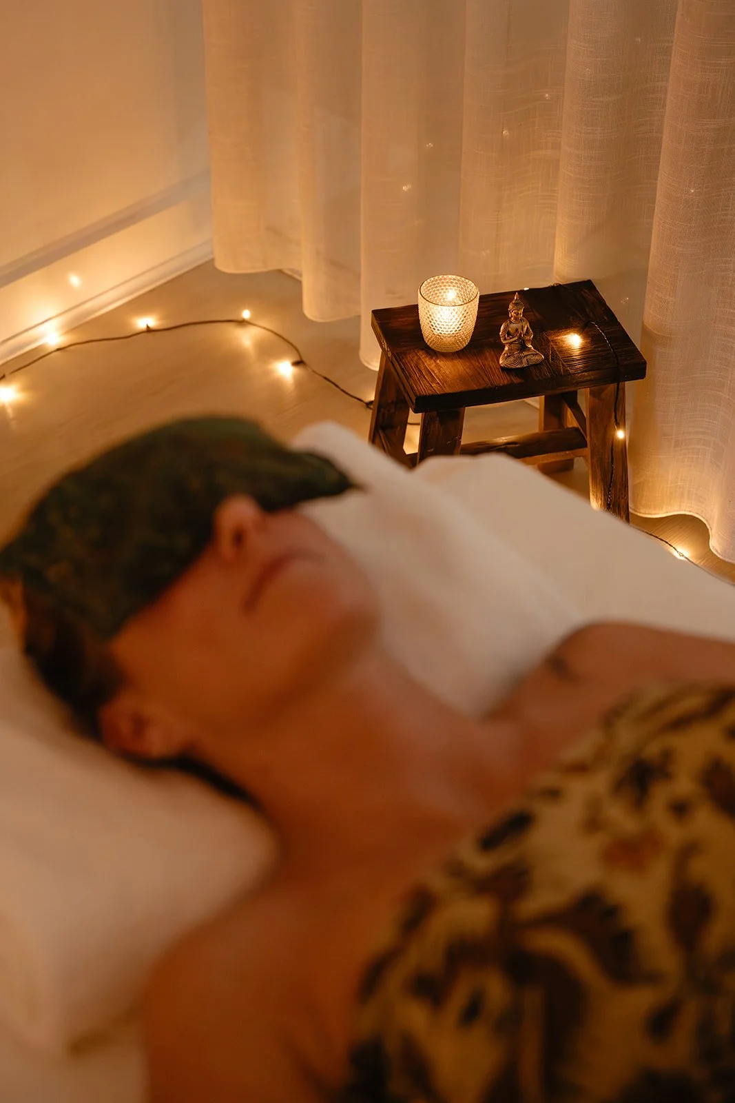A person sleeping on a bed with a wooden side table beside them. The table has a lit candle, a Buddha figurine, and a string of fairy lights. The background includes a sheer curtain and a string of fairy lights on the wall.