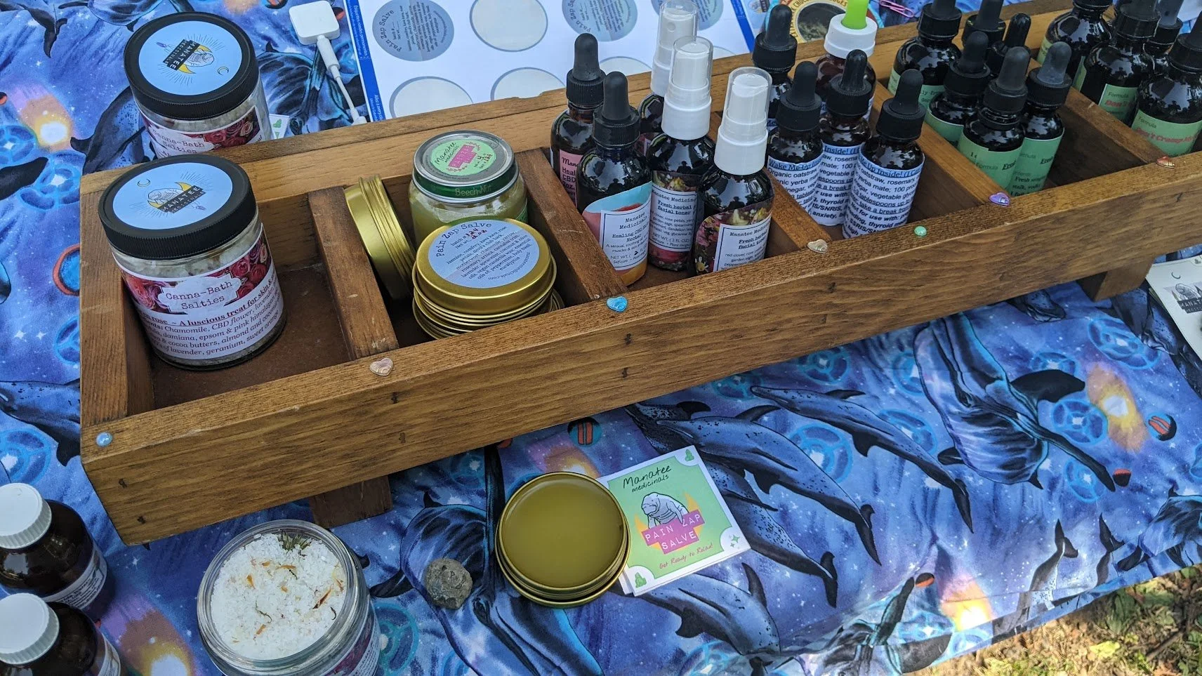 A close-up of Tavi's vendor table at an event, with tinctures, bath salts and salves displayed over a trippy blue dolphin fabric tablecloth