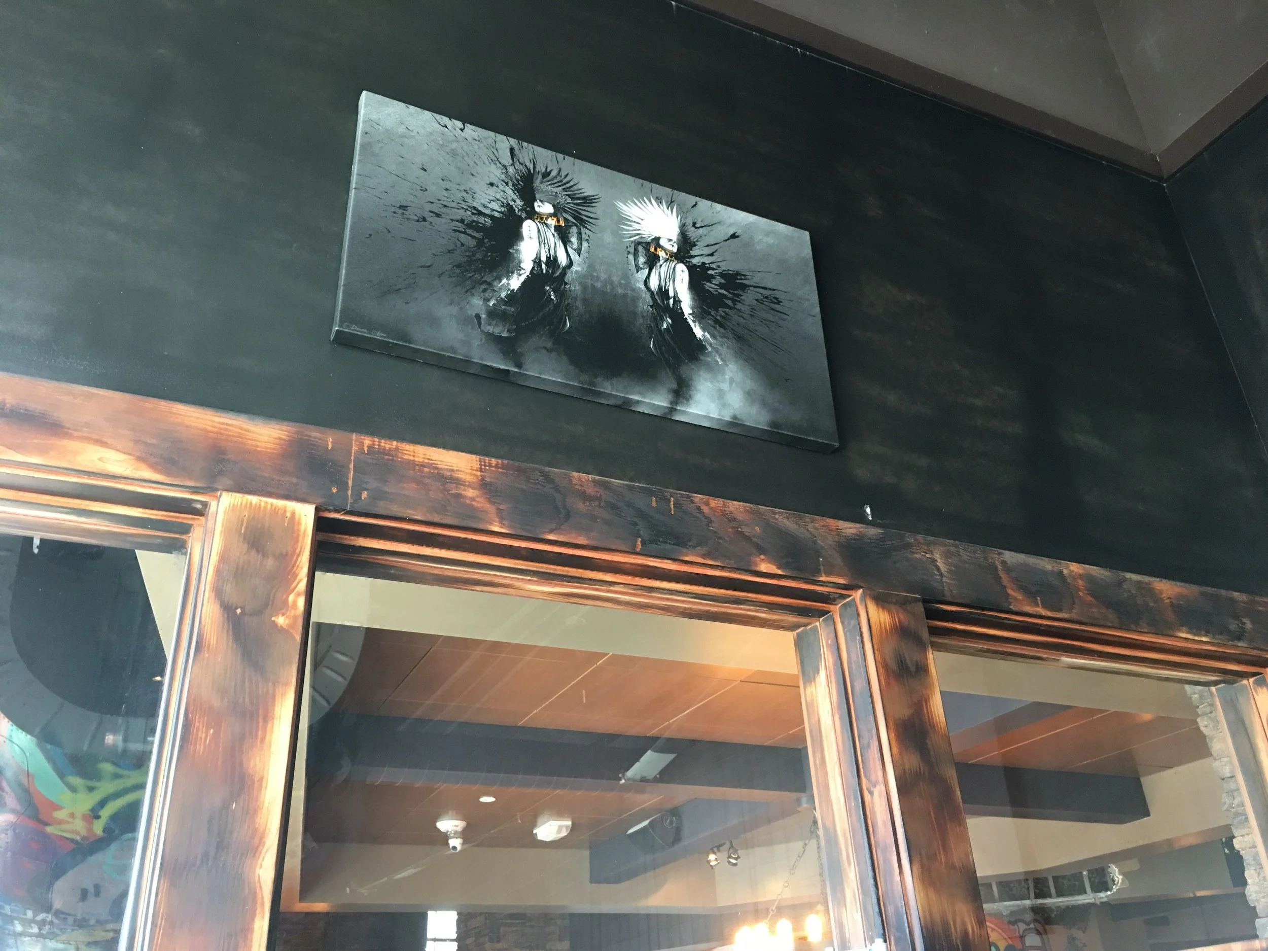 Black wall with a monochrome painting of two dancers wearing feathered headdresses, behind glass doors with wooden frames, in a modern interior space.