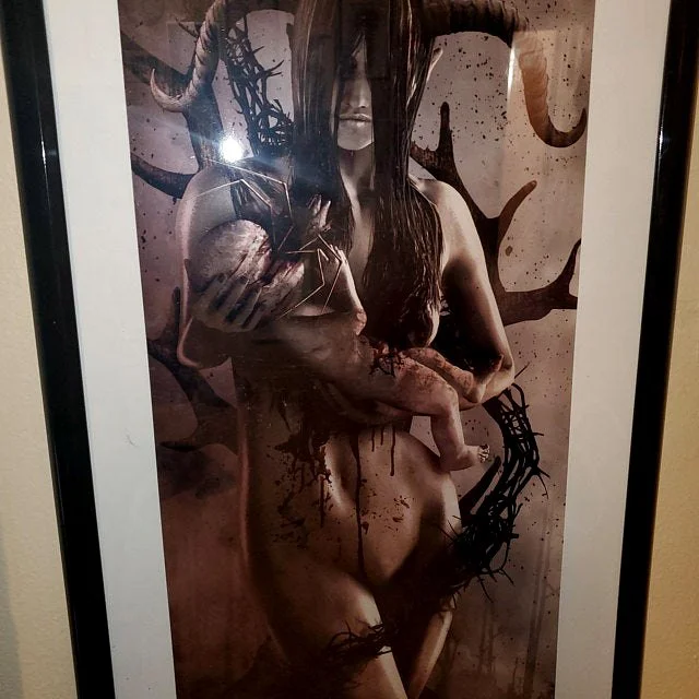 A dark, surreal artwork featuring a female figure with long hair, holding a baseball and a small, unconscious baby in her arms. The background and parts of her body are decorated with black, branch-like designs and splatters.