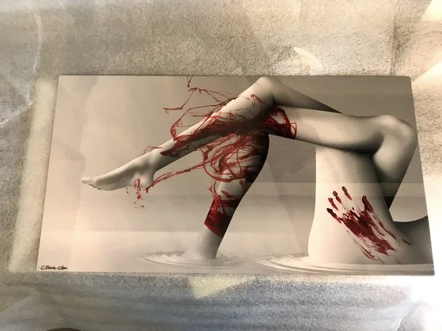 A black and white photo of a woman with red paint looking like blood, lying on the ground, with her head resting on her arm and her hair spread out. The woman is partially covered by a piece of paper or fabric, and there is red paint on her hand and 