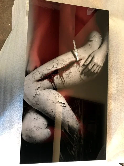 Artwork depicting a cracked, weathered sculpture of a human leg with a knife stabbing into the thigh and simulated blood.