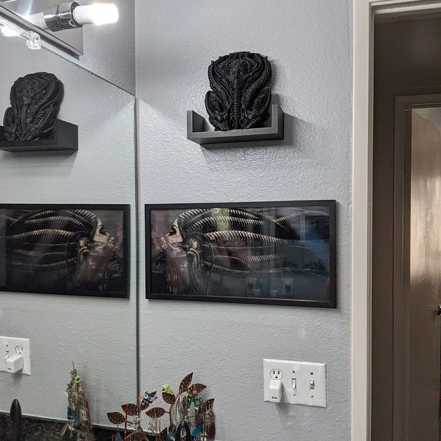 Black decorative sculpture on a wall shelf above framed picture and electrical outlets in a room.