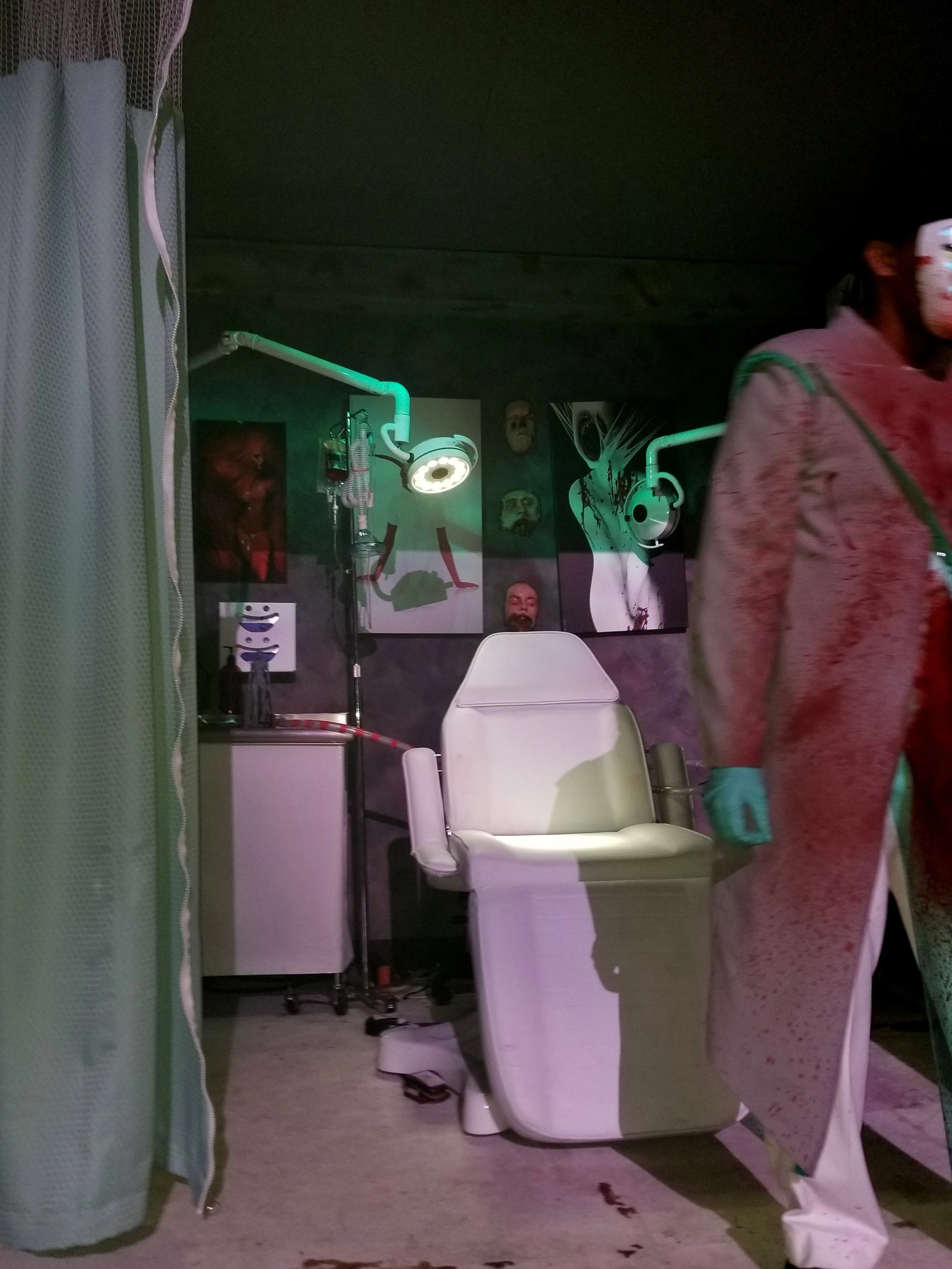 Medical examination chair in a dimly lit room with dark walls decorated with horror-themed artwork and masks, medical equipment, and a person wearing medical gloves and a face mask partially visible on the right side.