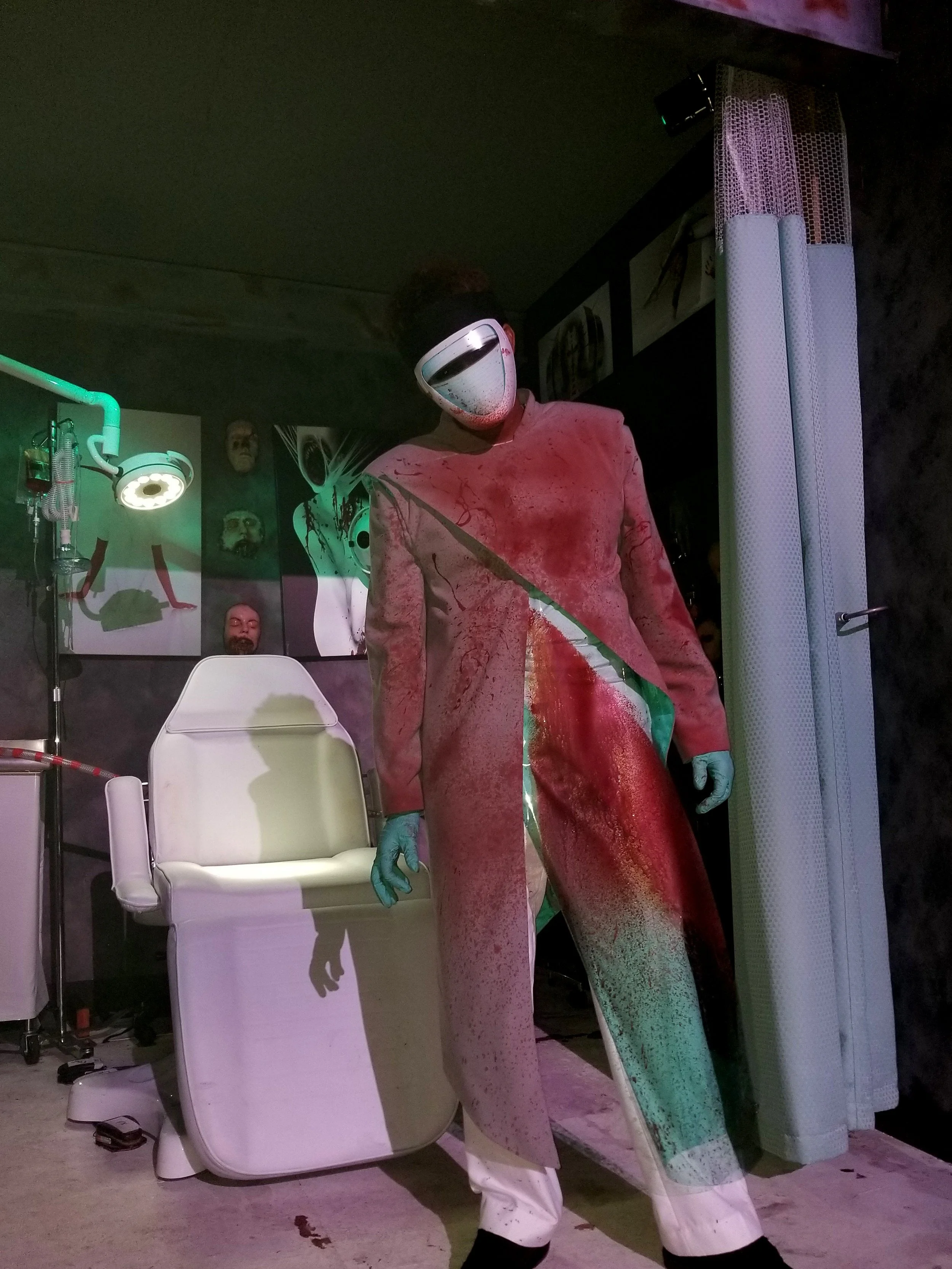 A person dressed as a surgeon or scientist with a white visor mask, blue gloves, and a long dirty coat standing in a laboratory or medical setting.