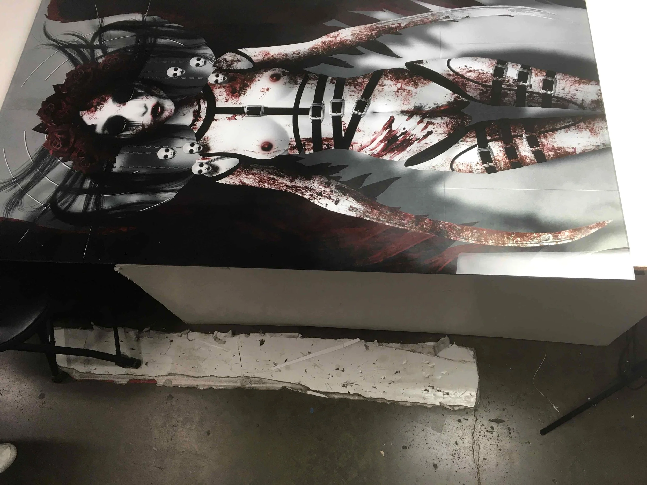 A large, dark artwork of a woman with black hair, blood splatters, black horns, and an eyepatch, with multiple eyeball necklaces. The image is printed on a surface that is slightly damaged at the bottom.