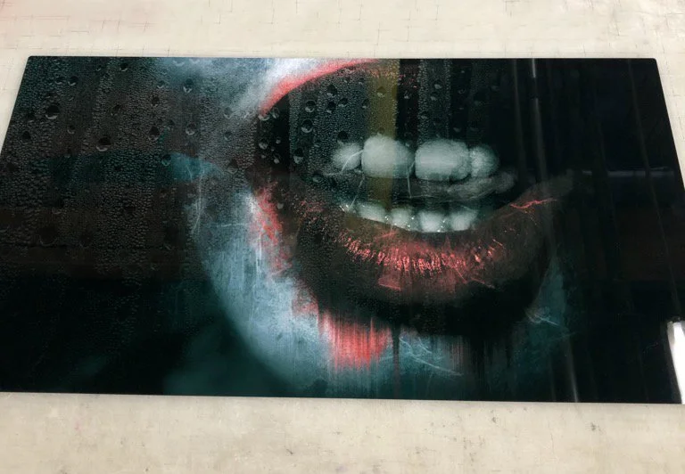 Close-up of a person's lips painted with metallic red lipstick, partially open revealing teeth, with a water droplet-covered surface in the foreground.