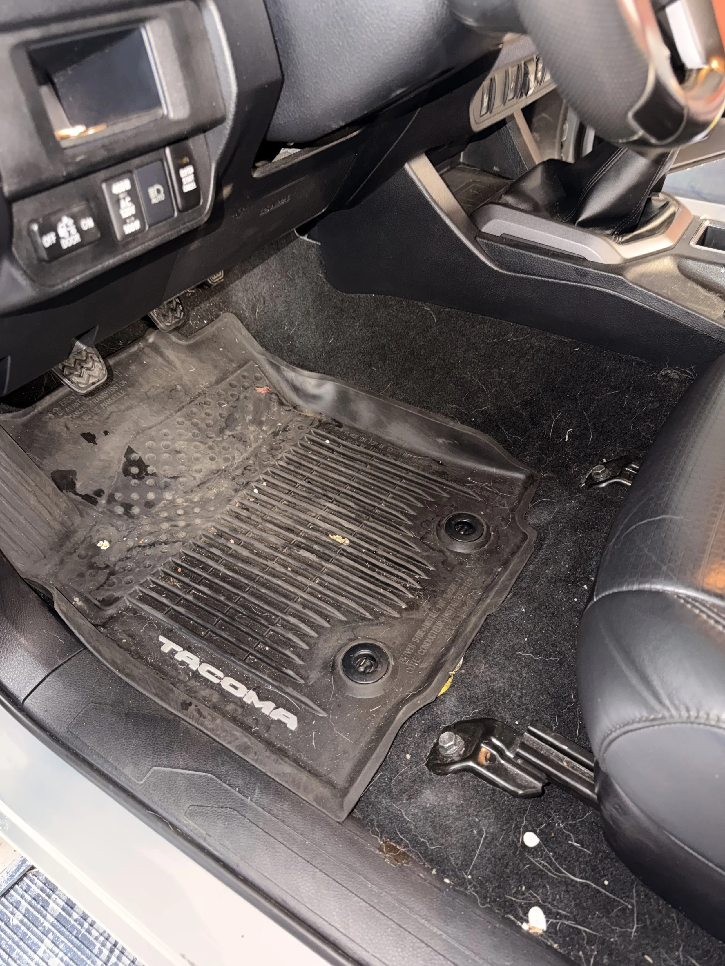 Inside of a vehicle showing a dirt and debris-covered floor mat with the Toyota Tacoma logo, adjacent to the driver's seat and gear shift.